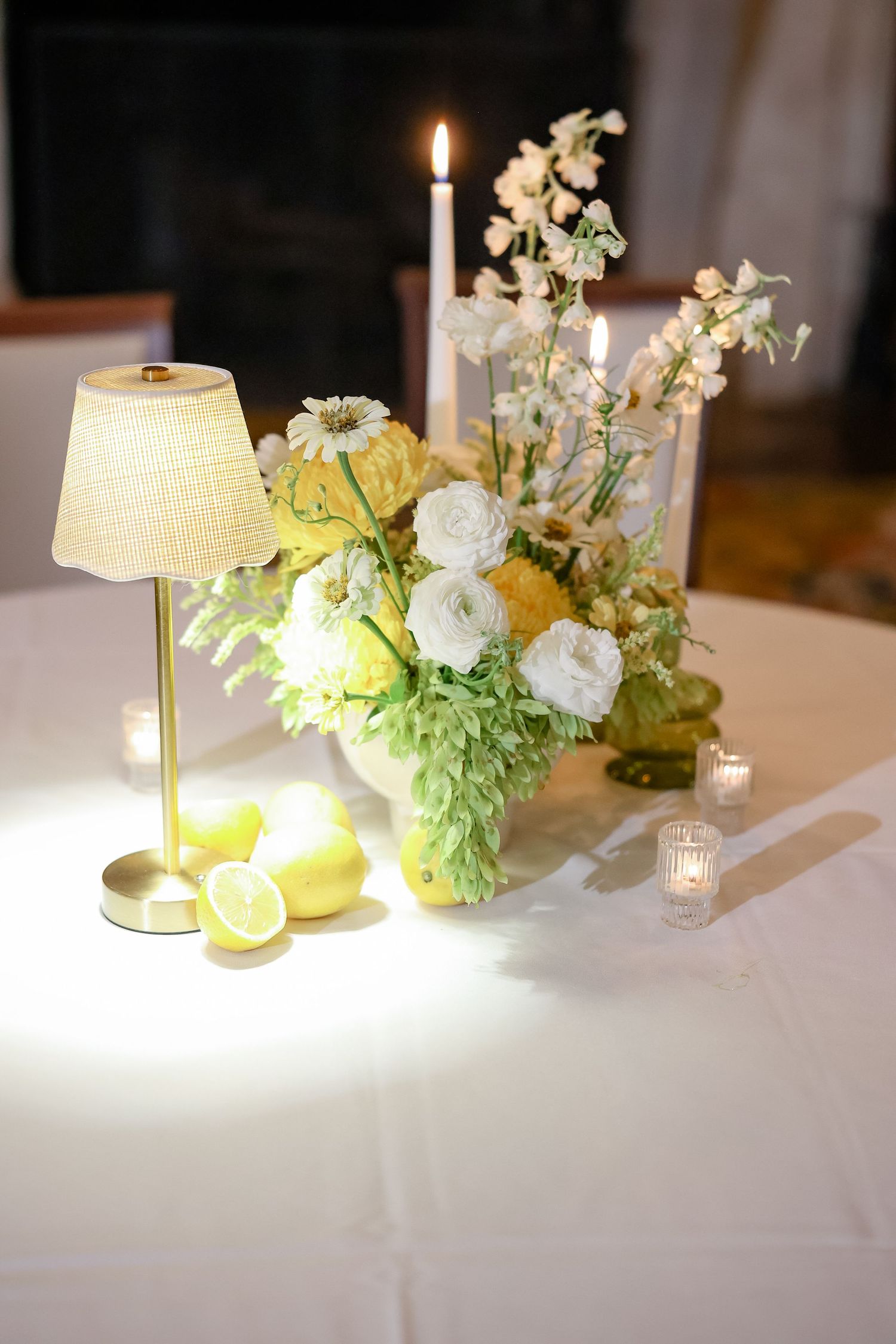 Wedding table centerpiece with white and yellow flowers, lemons, a small lamp, and candles on a white linen.