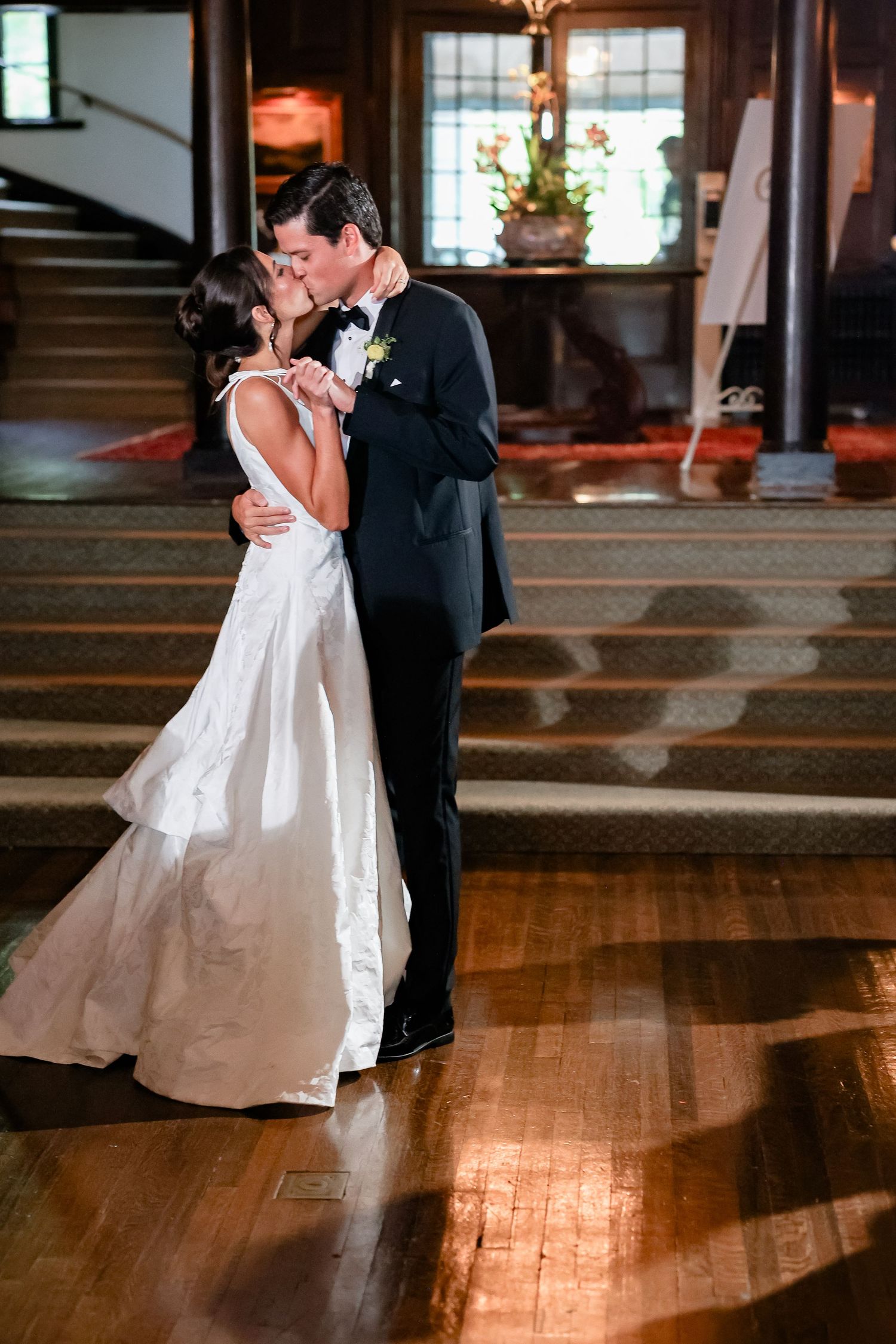 Bride and groom share first dance kiss in elegant venue with hardwood floors and grand staircase.