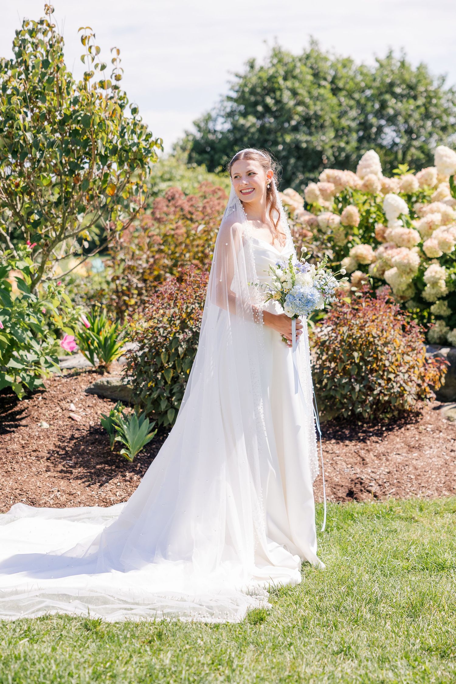 Bride in white gown and long veil holding blue bouquet standing in lush garden on wedding day.