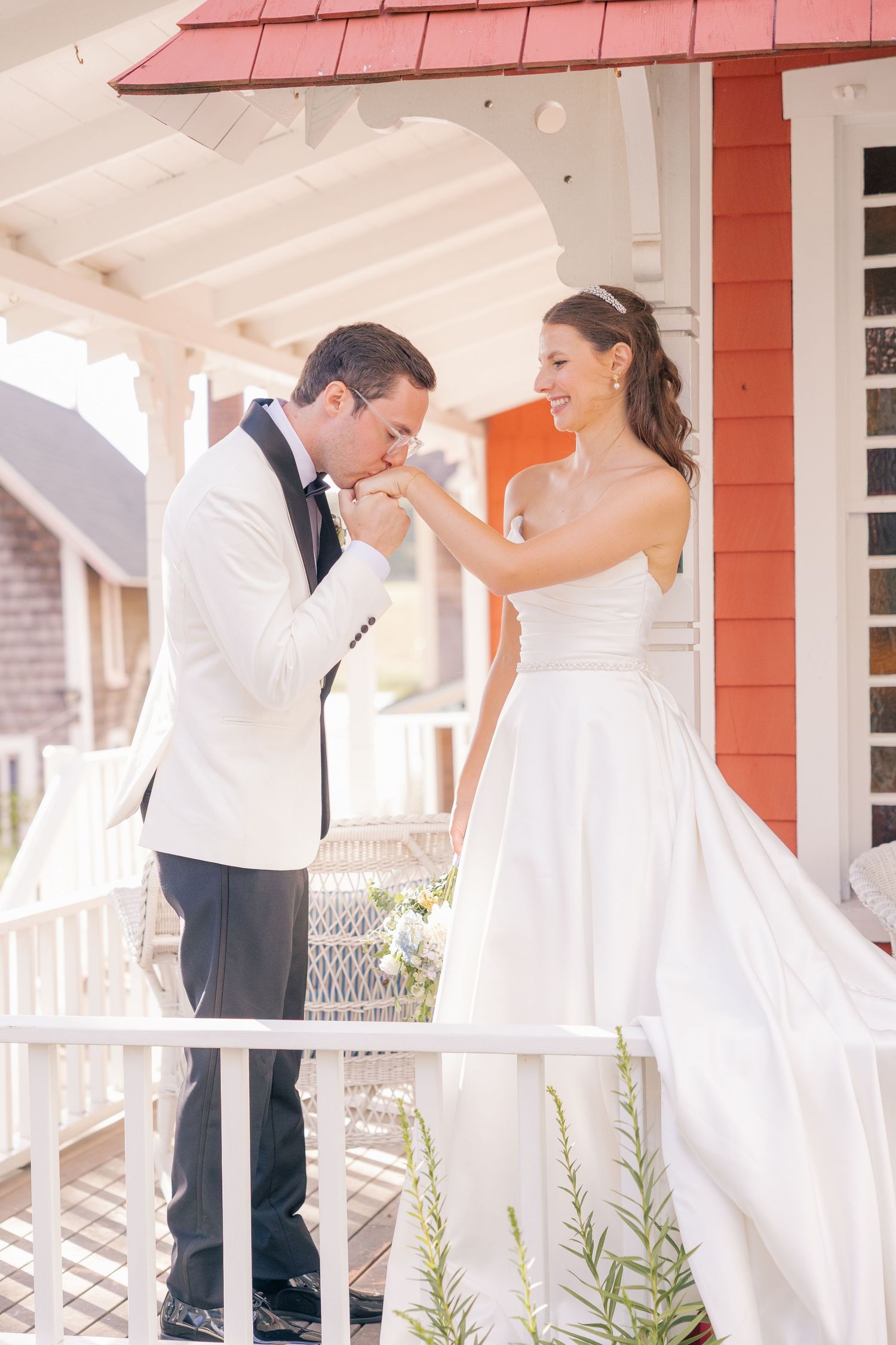 Groom in white tuxedo kissing bride's hand on porch, bride in strapless white gown smiling joyfully.
