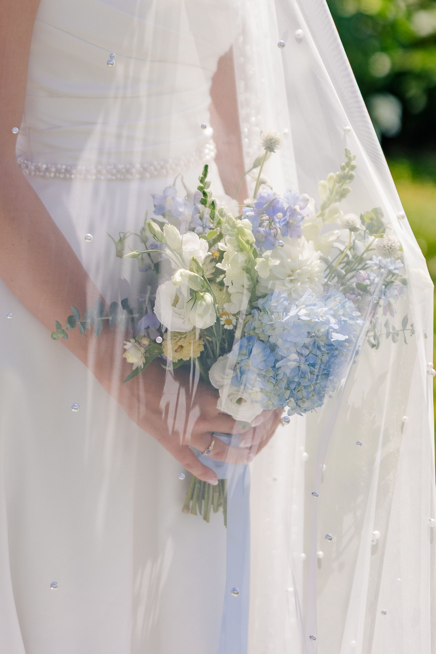 Bride holding a delicate blue and white wedding bouquet with roses and delphiniums, draped under a pearl-embellished veil.