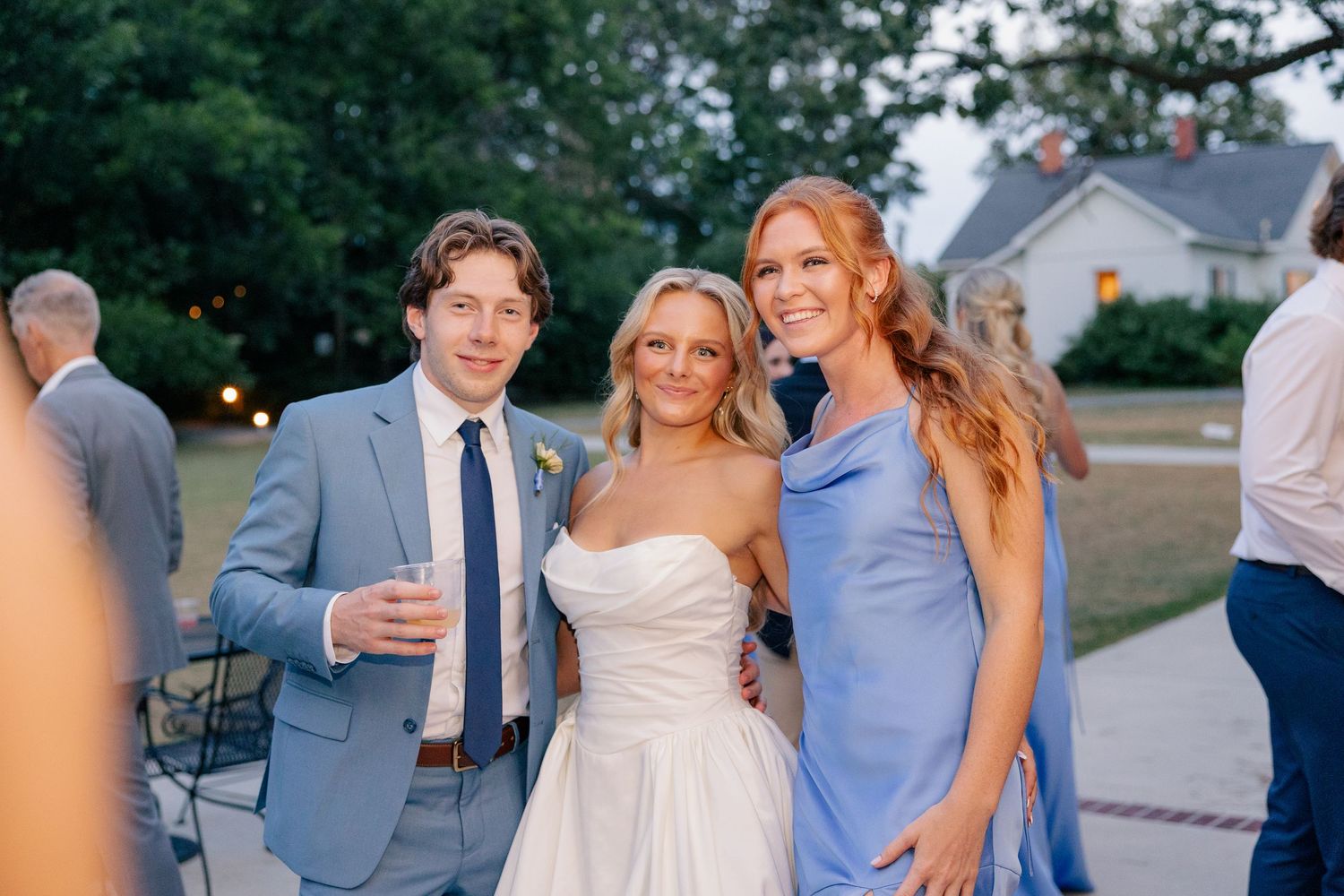 Bride in strapless white gown poses with groom in blue suit and bridesmaid in blue dress at outdoor wedding reception.