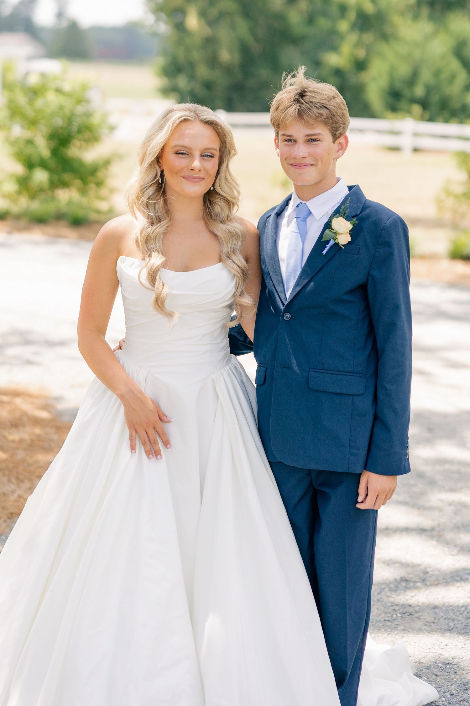 Bride in white strapless ball gown poses with young man in navy suit and boutonniere outdoors.