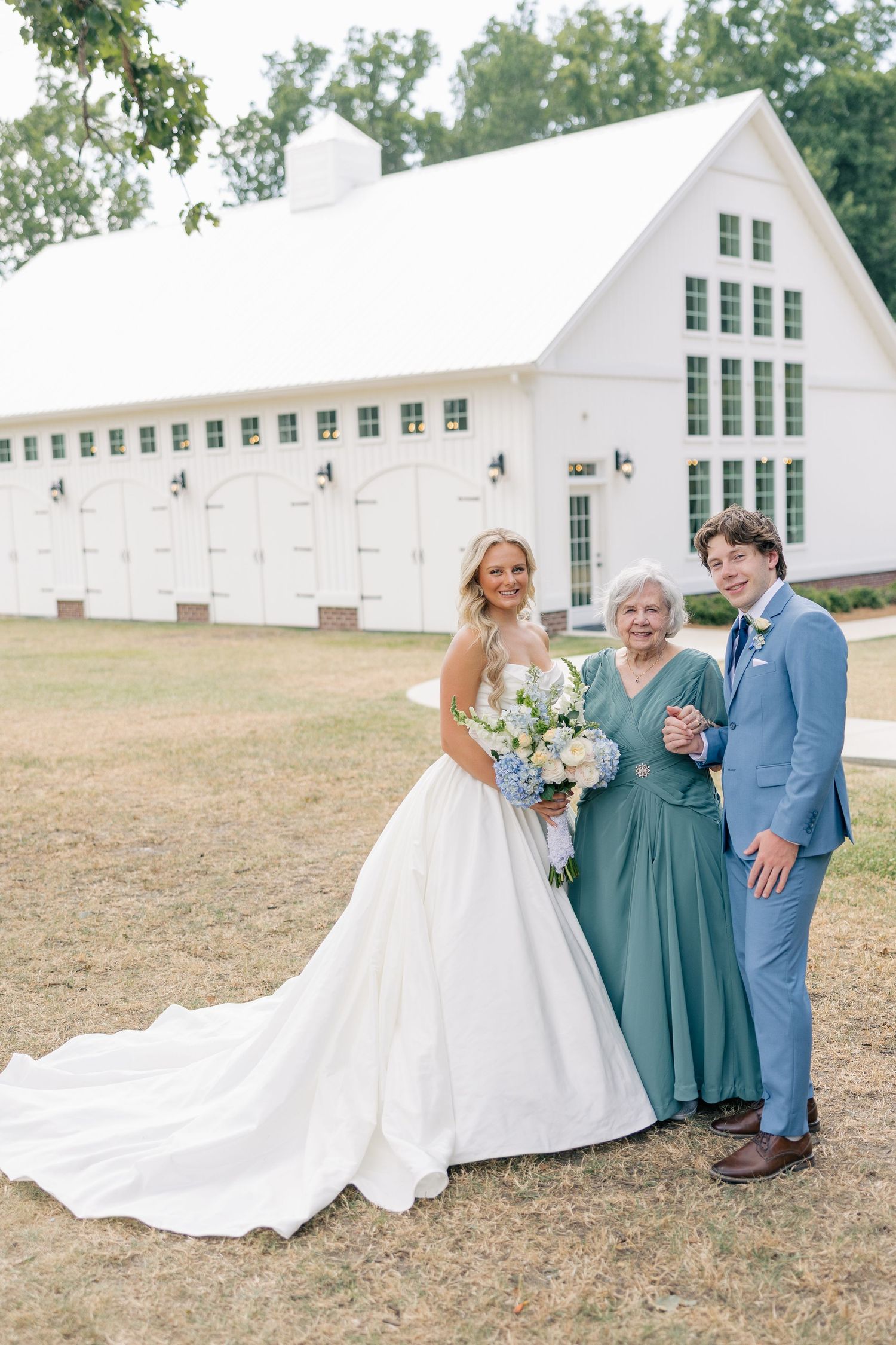 Bride in white ball gown holds bouquet with groom in blue suit and elderly woman in teal dress outside white barn.