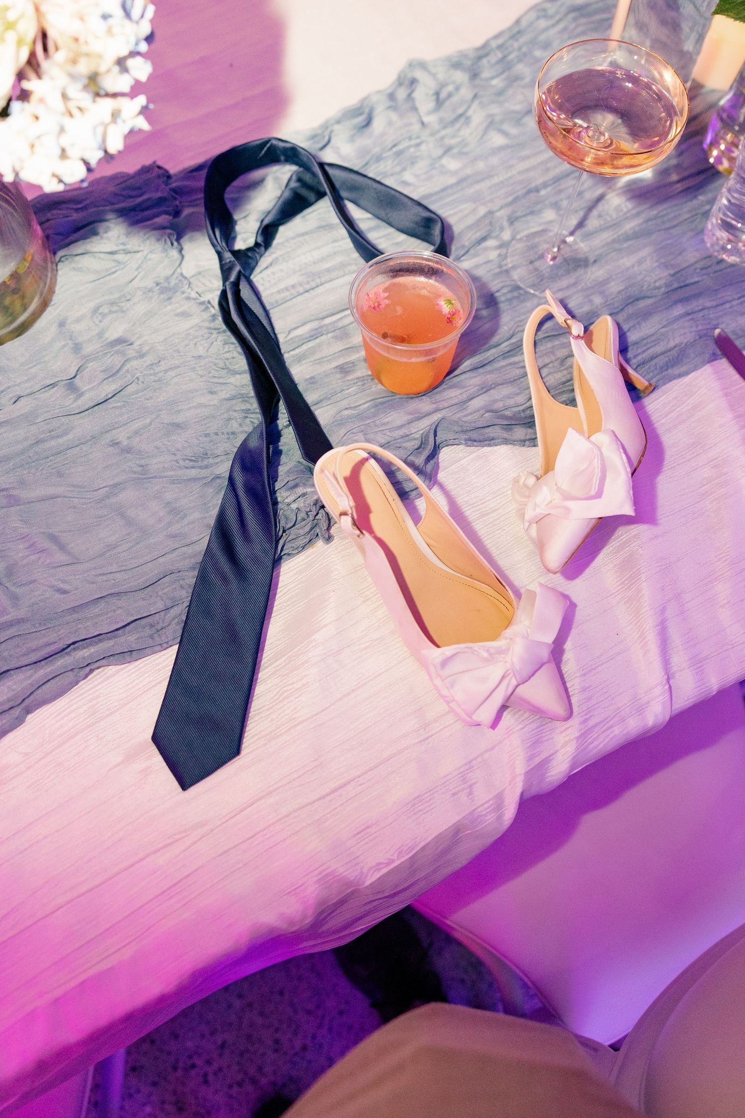 Bridal details flatlay featuring white bow heels, black tie, and rose drink on a draped table.
