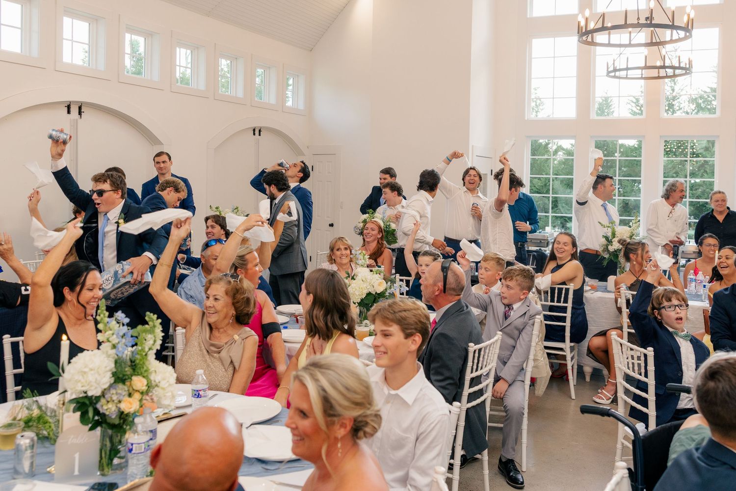 Wedding guests celebrate with raised hands and napkins during lively reception in elegant white venue.
