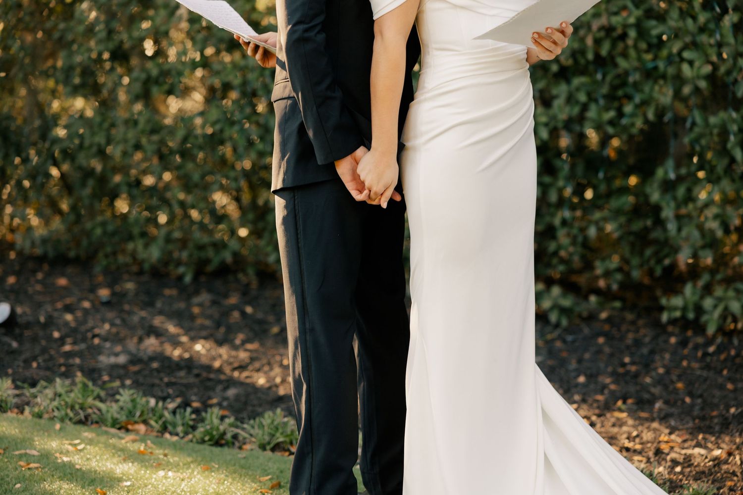 Couple holds hands tenderly during outdoor wedding ceremony wearing black suit and white gown.