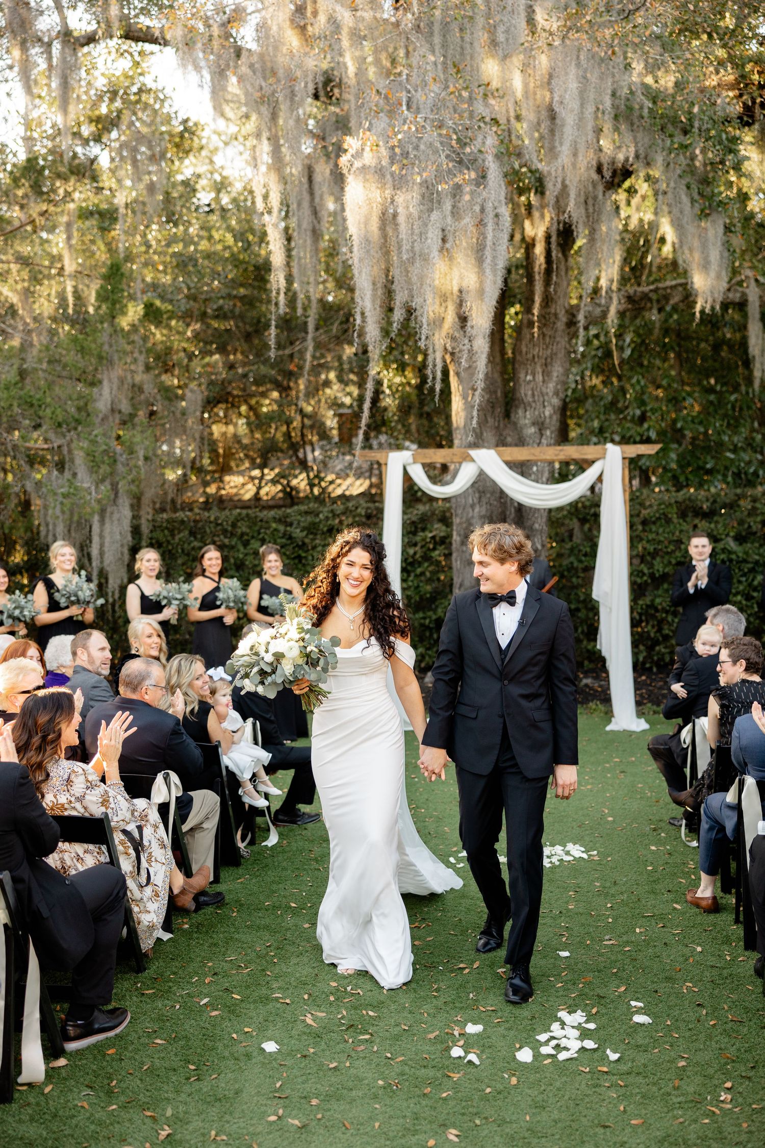A couple walks down the aisle at their outdoor ceremony under Spanish moss draped trees and white arch decorations.