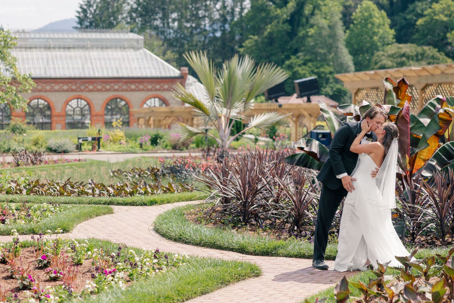 Bride and groom share a romantic dip kiss in lush garden with greenhouse backdrop.