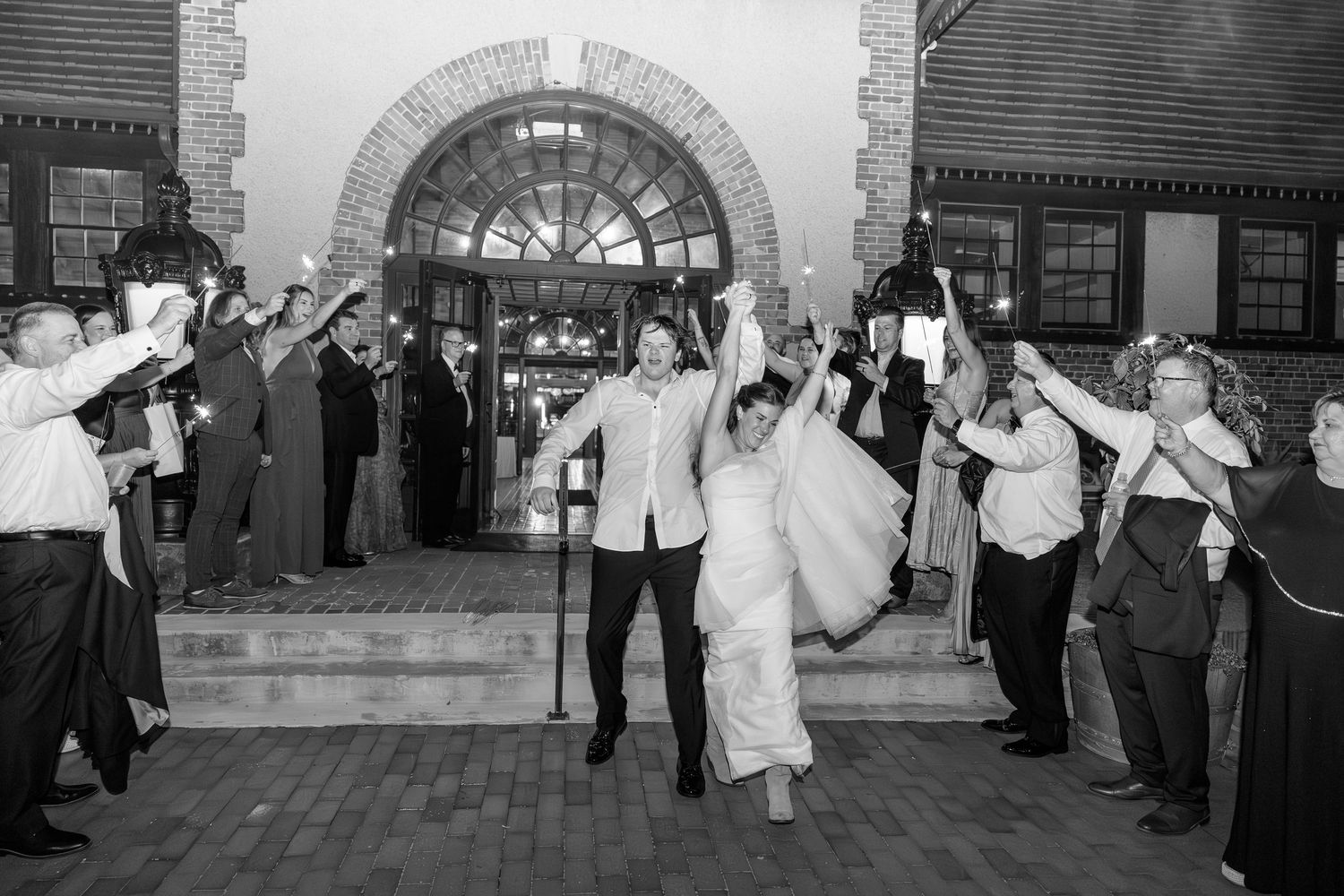 Newlyweds exit ceremony under raised hands of cheering guests in black and white photo.