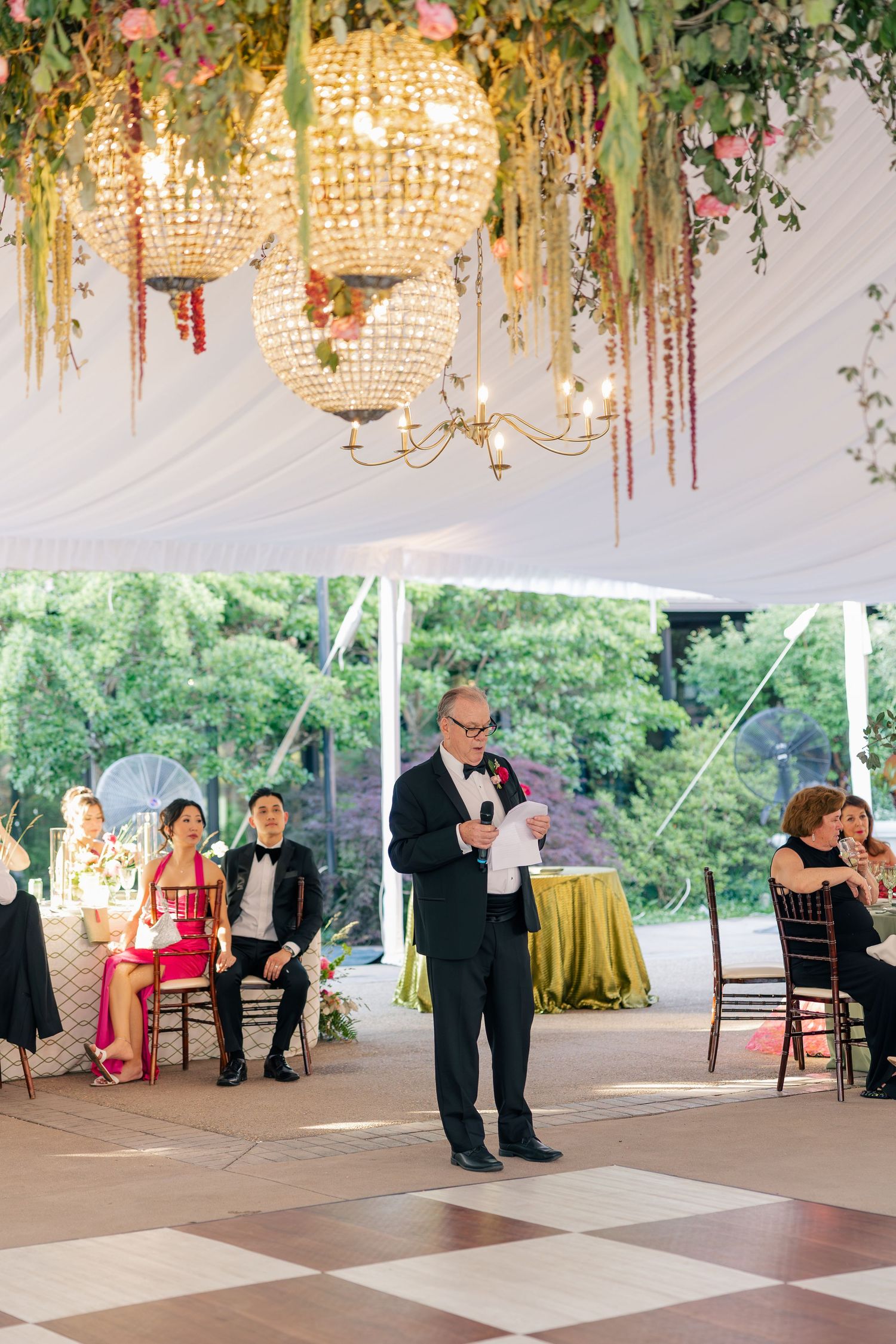 Father of the bride delivers heartfelt speech on checkered dance floor beneath floral chandeliers at reception.