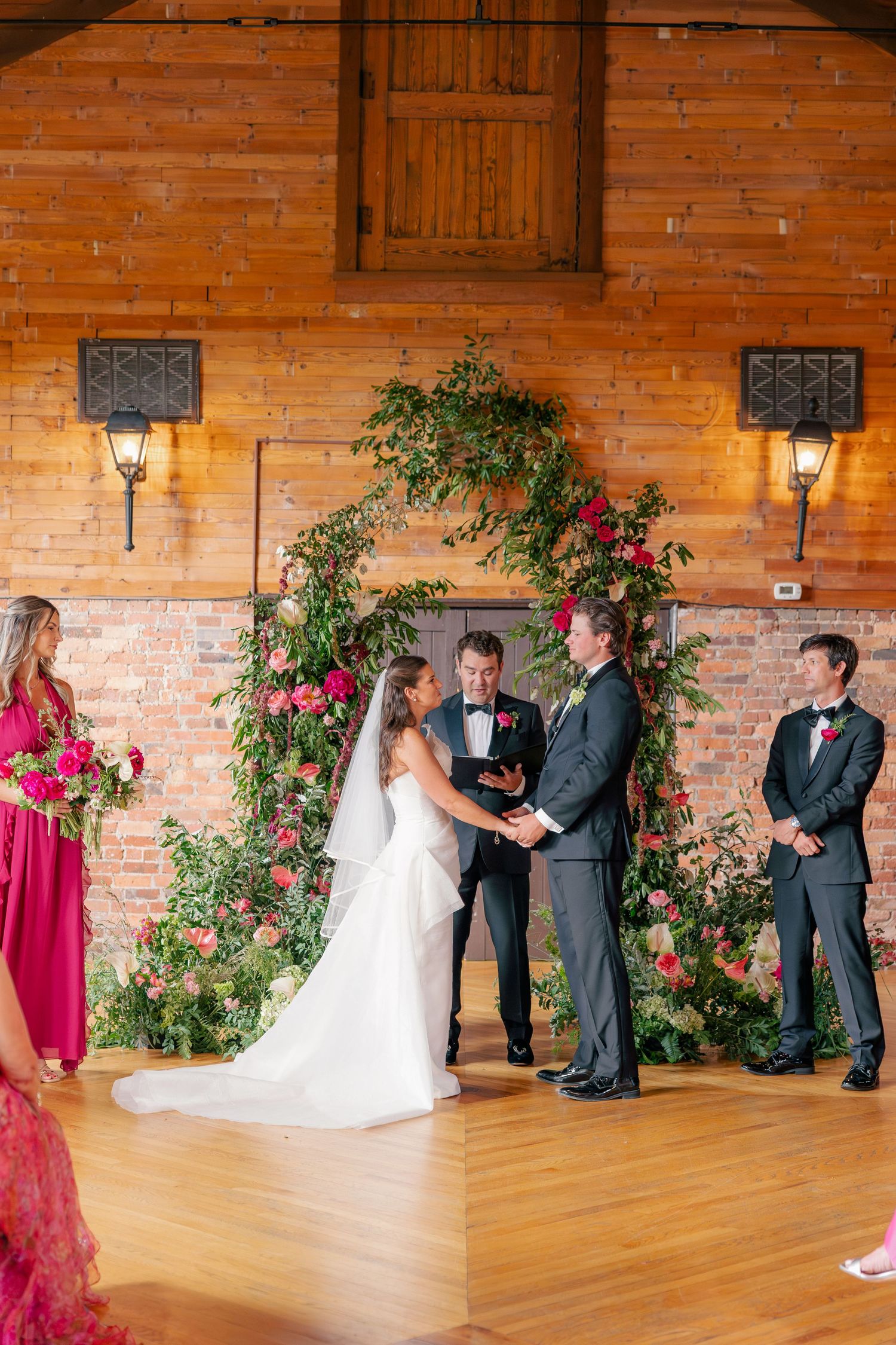 Bride and groom exchange vows at lush greenery and pink floral arch in elegant barn ceremony.