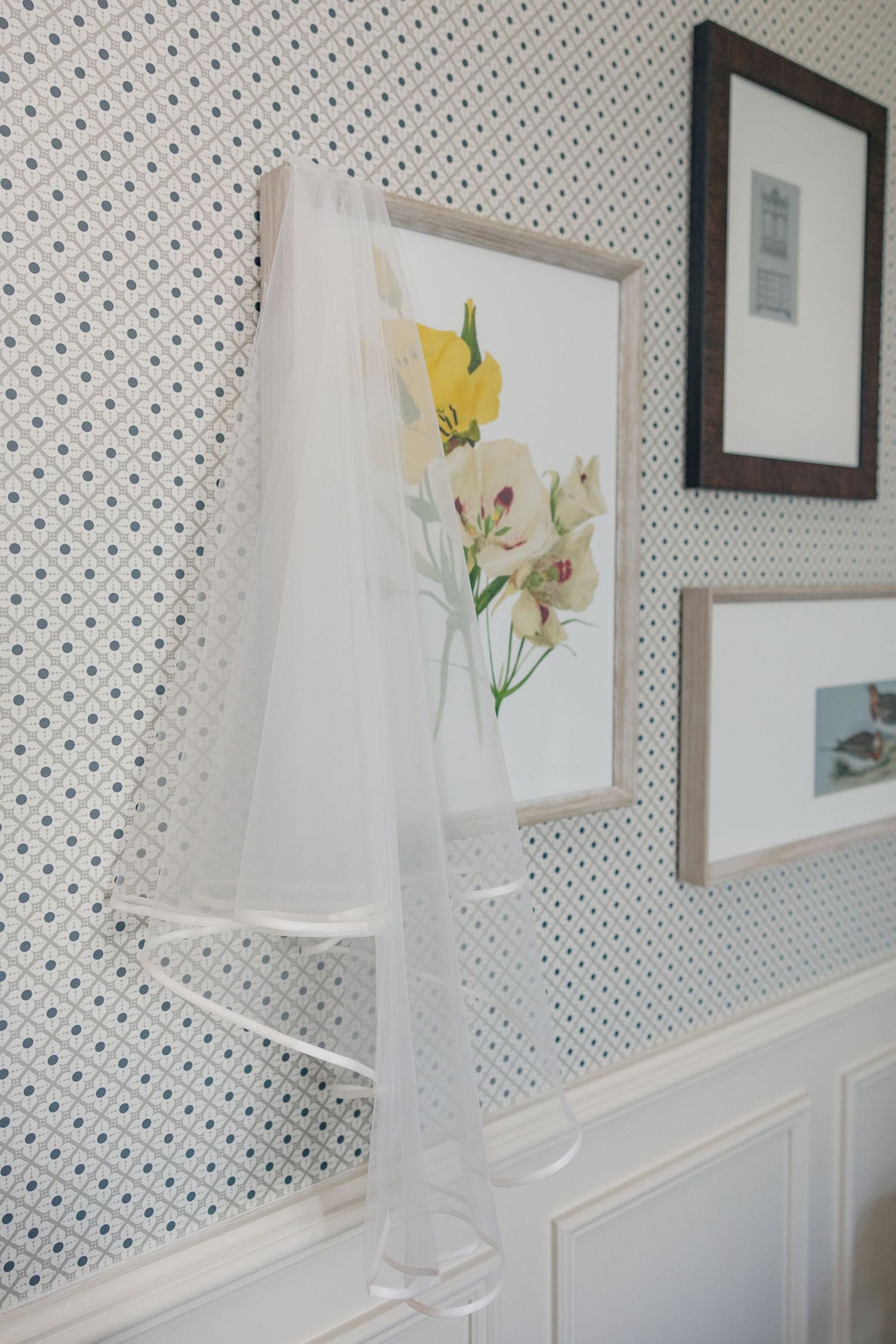 A delicate white bridal veil hangs on a wall beside floral botanical artwork in a room with patterned wallpaper.