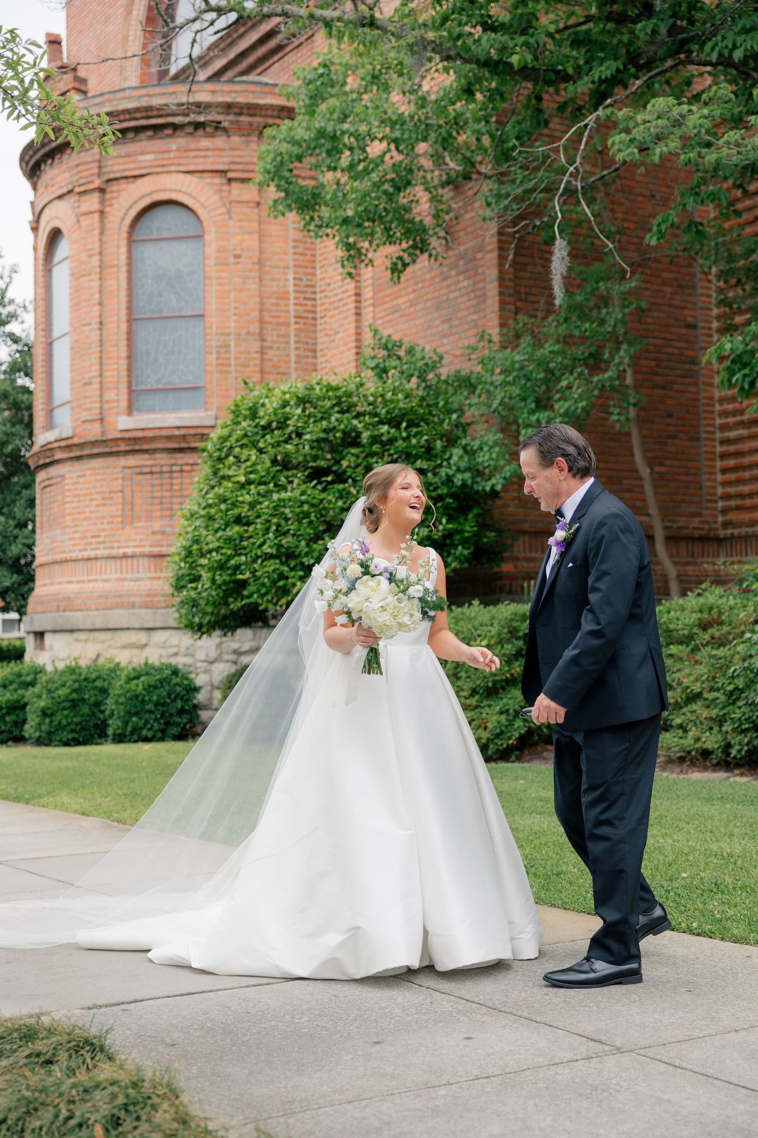 Bride laughing joyfully with her father during first look outside a historic red brick church.