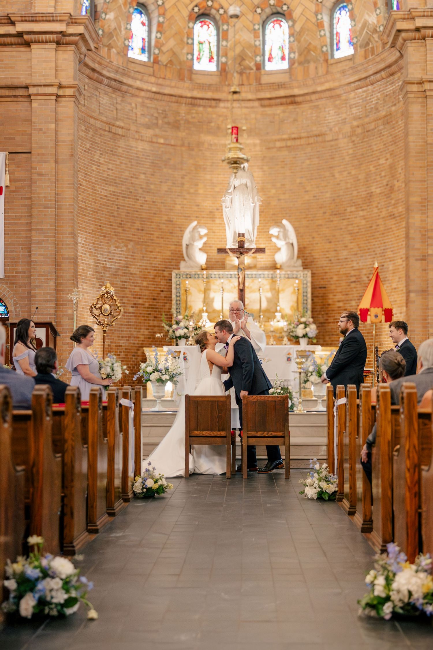 Bride and groom share first kiss at ornate Catholic church altar during wedding ceremony with floral arrangements lining the aisle.