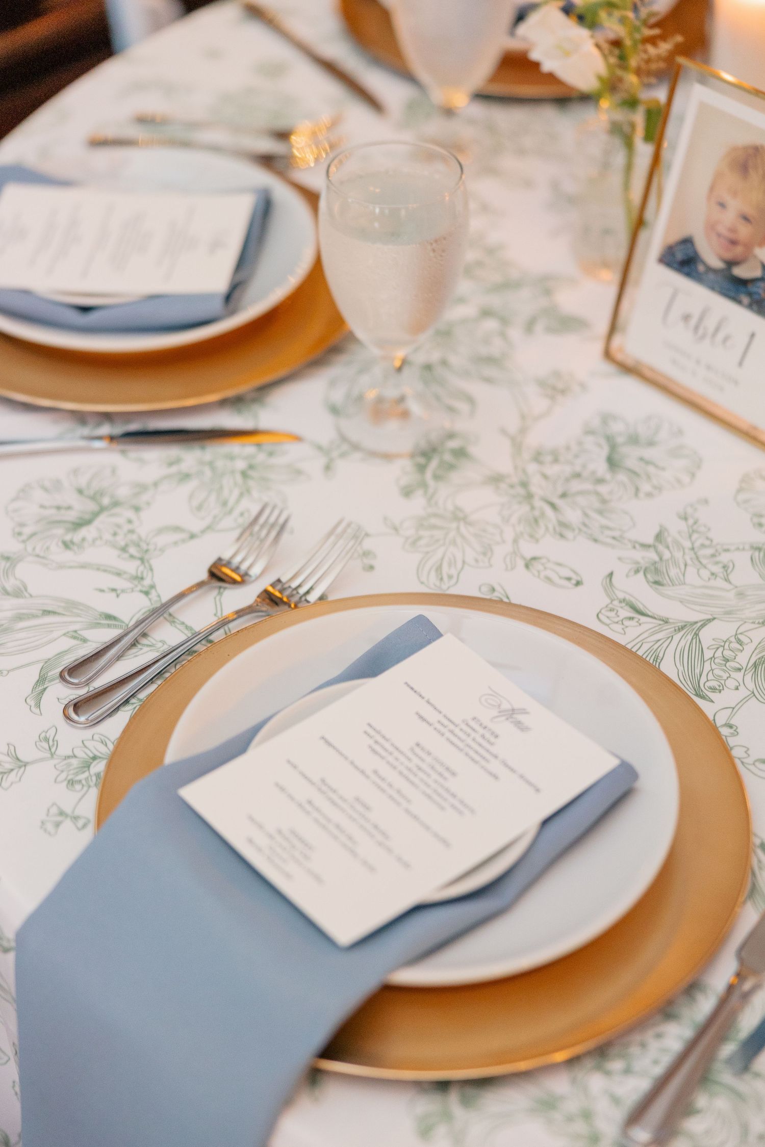 Elegant wedding place setting with gold charger, blue napkin, menu card, and floral green tablecloth.