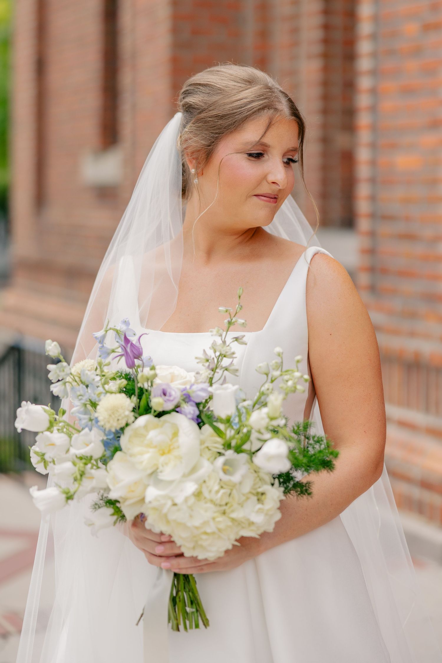 Bride in white square-neck gown holding lush white and lavender bouquet with veil outdoors.