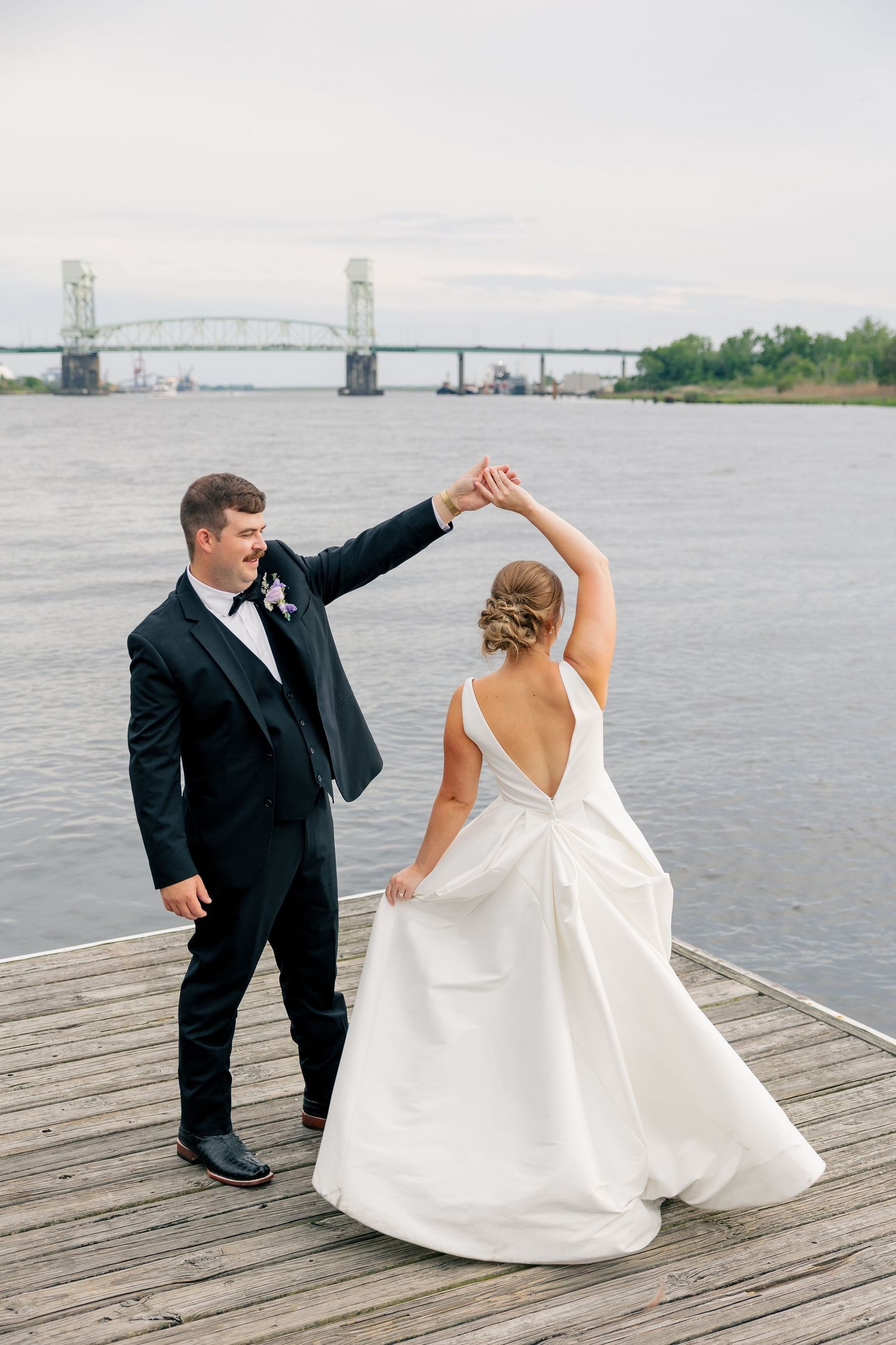 Bride and groom dance on wooden dock with scenic river bridge backdrop on their wedding day.