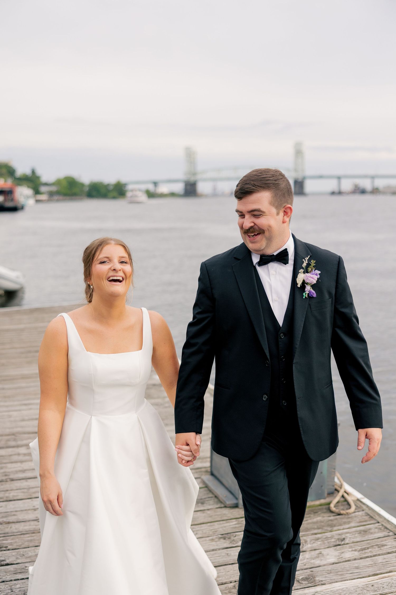 Laughing bride and groom walk hand-in-hand on a waterfront dock with a bridge in the background.