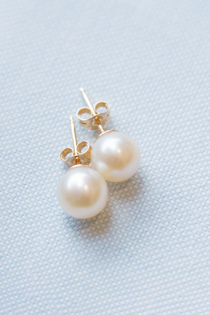 A pair of classic white pearl stud earrings with gold settings on a light blue fabric background.