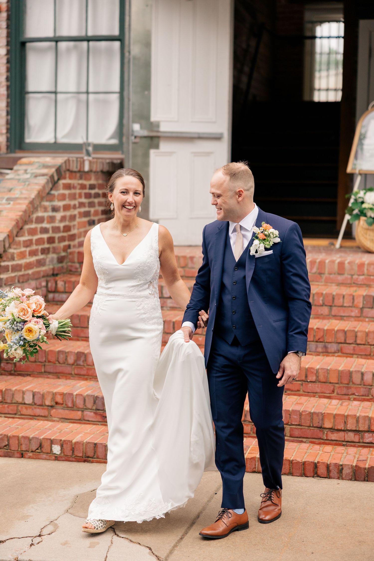 Joyful newlyweds walk hand-in-hand down brick steps after wedding ceremony, bride holding bouquet.
