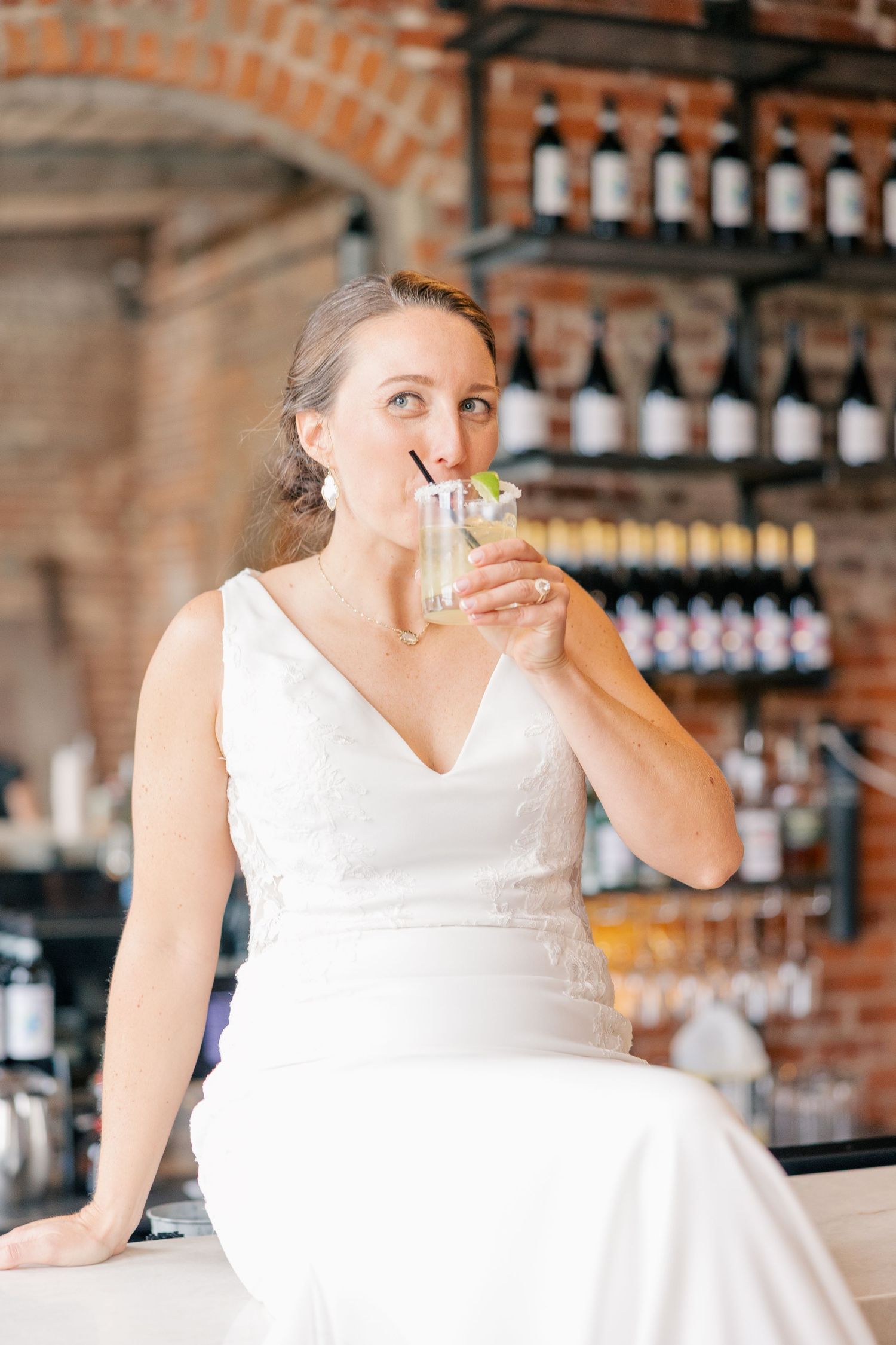 Happy bride in white gown sips cocktail while seated at rustic brick bar on wedding day.
