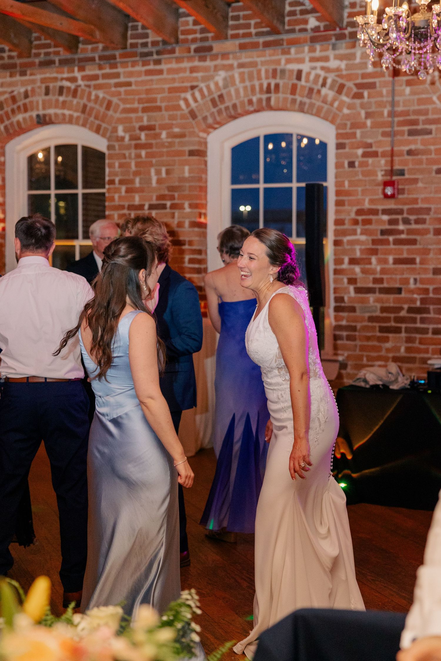 Women in elegant gowns dancing and socializing at a wedding reception in a rustic brick venue with chandelier lighting.