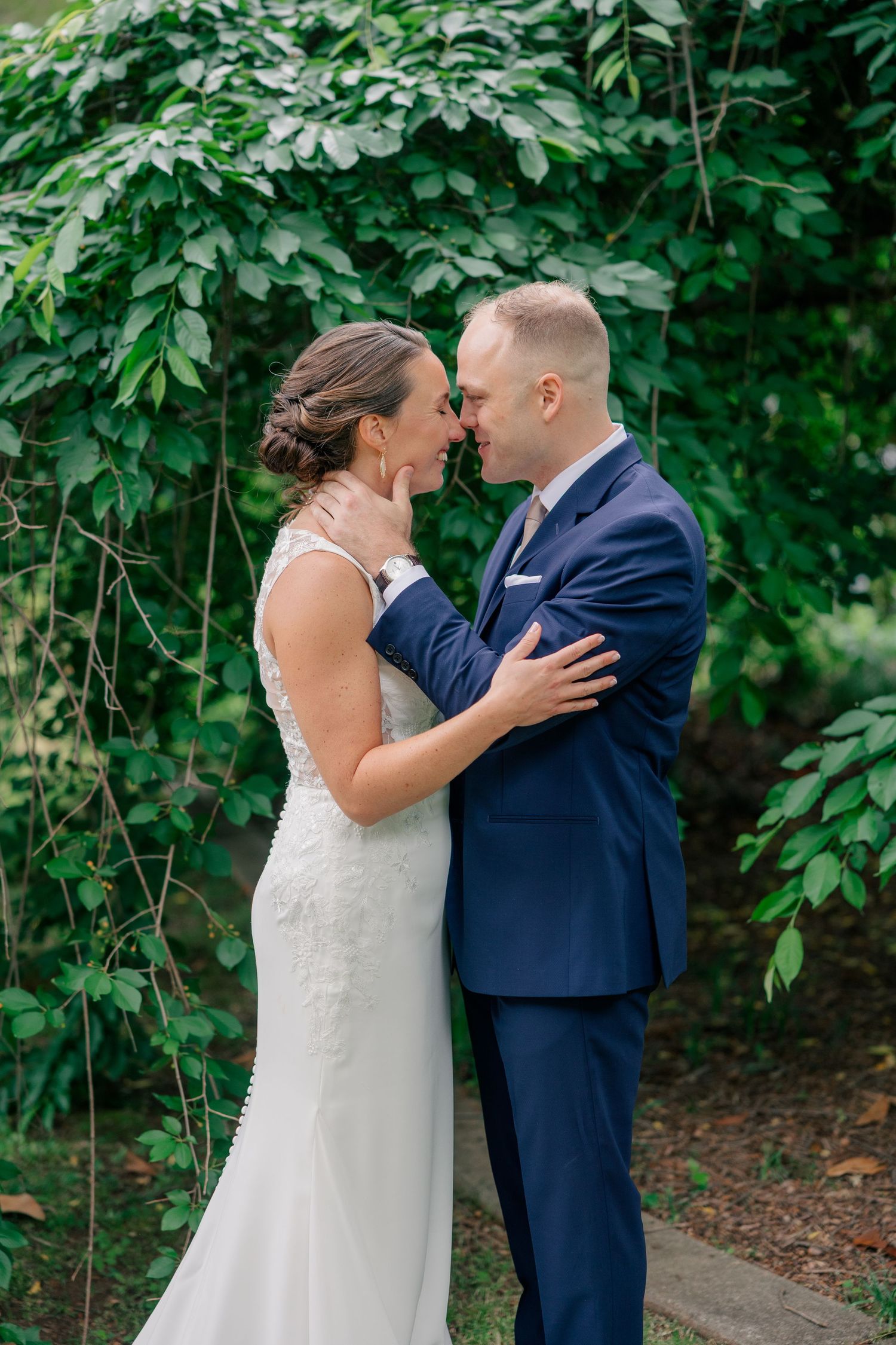 Bride and groom share romantic moment nose-to-nose surrounded by lush green foliage on wedding day.
