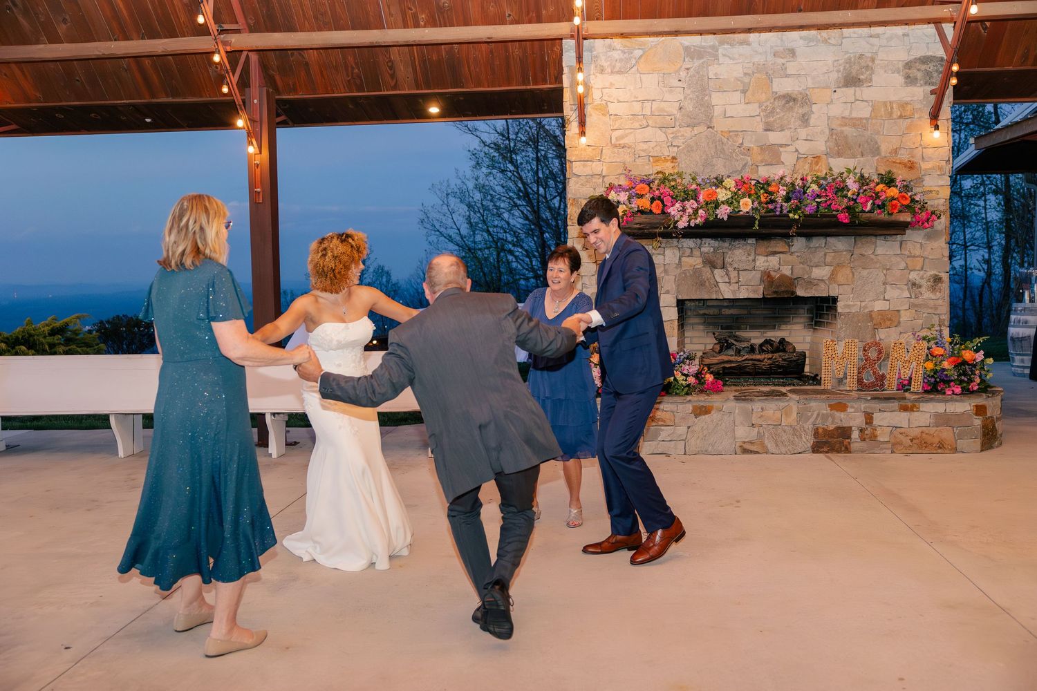 Wedding guests dance together on covered outdoor patio with stone fireplace and colorful floral decor.