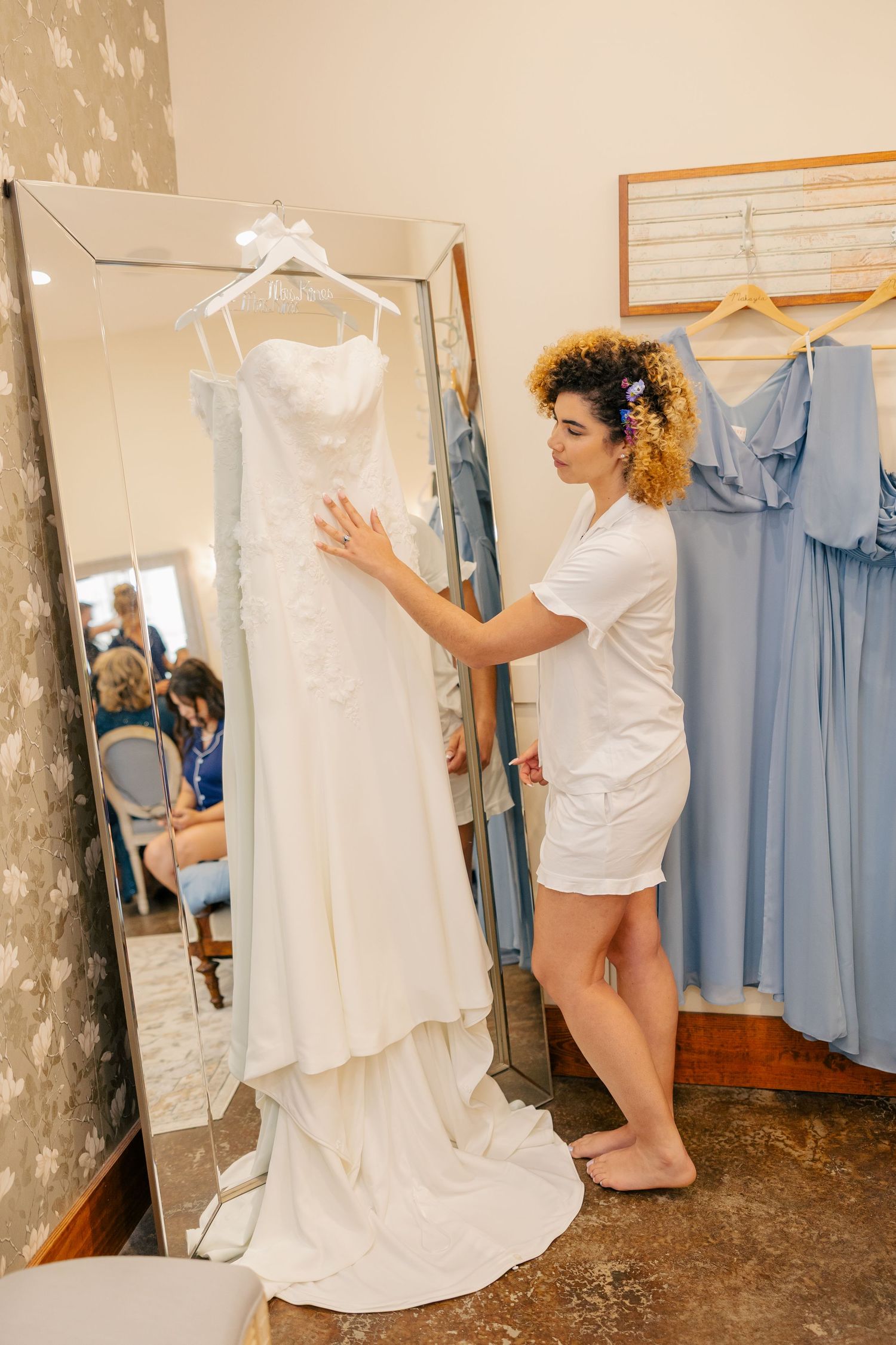 Bride examining white lace wedding gown on hanger next to mirror in bridal suite.