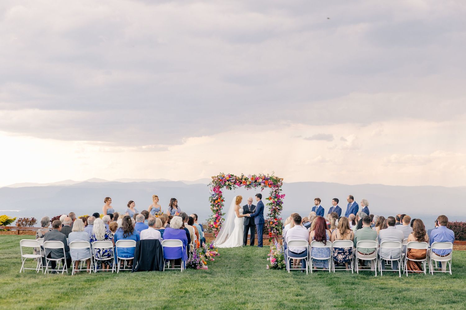 Outdoor wedding ceremony with guests seated on white chairs facing couple at colorful floral arch.