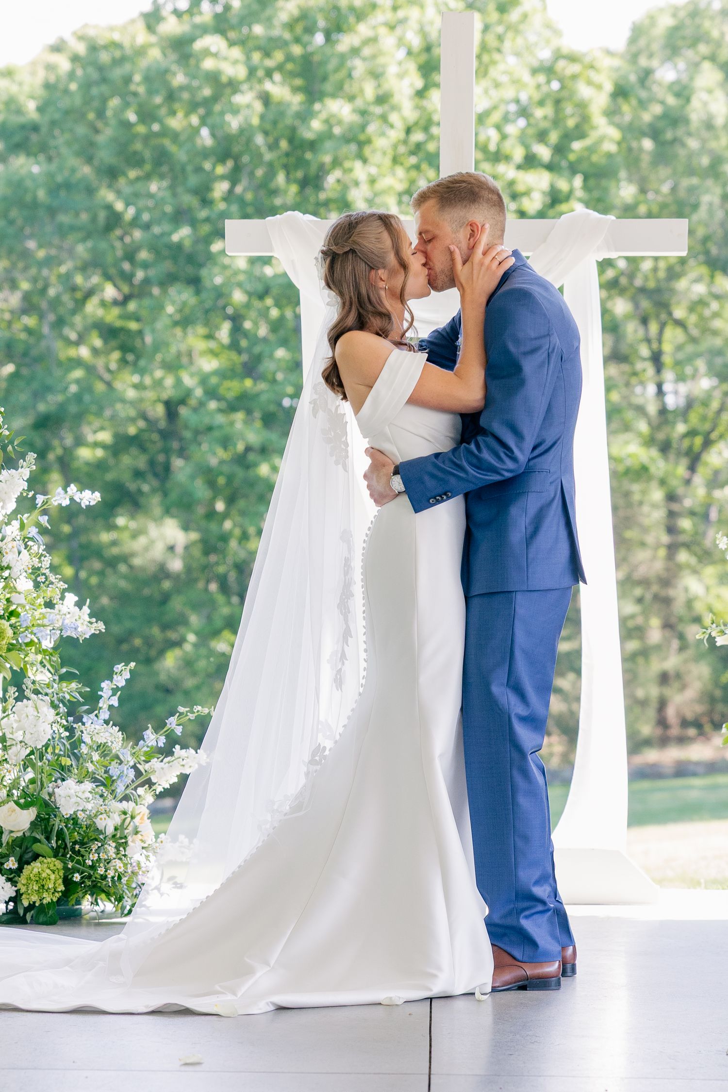 Bride and groom share first kiss before white cross altar surrounded by white florals.