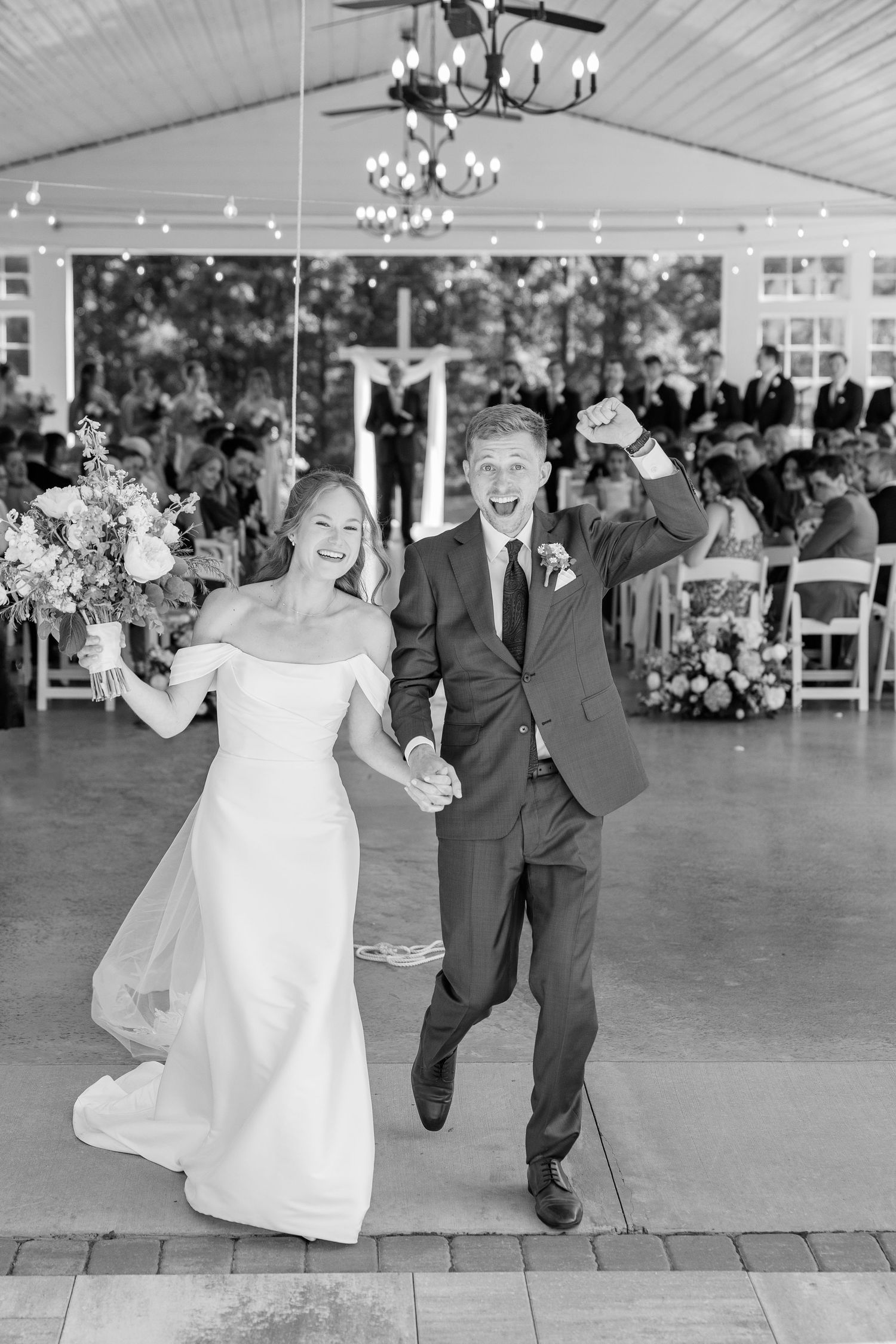 Black and white photo of joyful newlyweds celebrating with raised fists while exiting wedding ceremony pavilion.