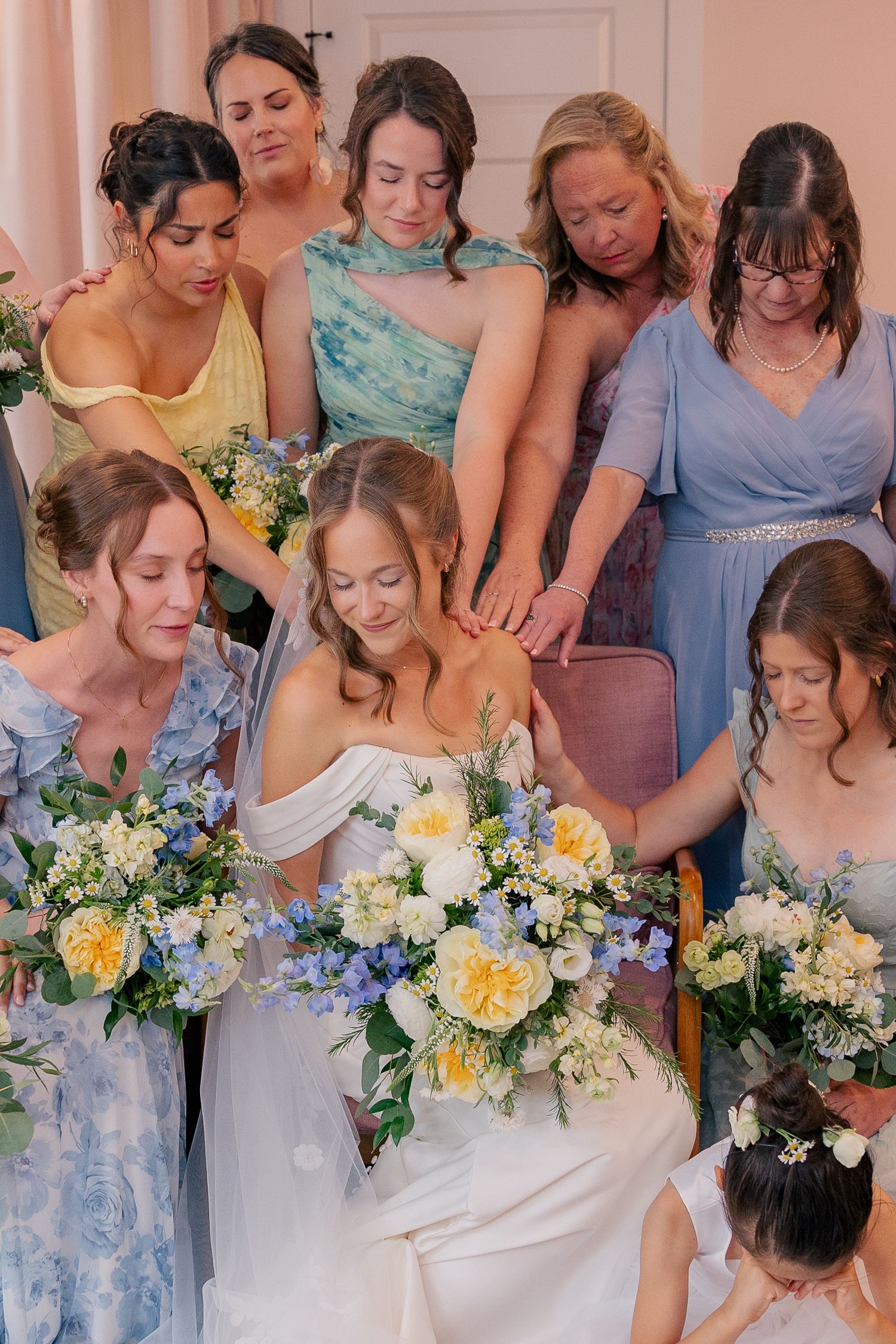 Bride surrounded by bridesmaids in blue and yellow dresses holding floral bouquets during a wedding prayer moment.