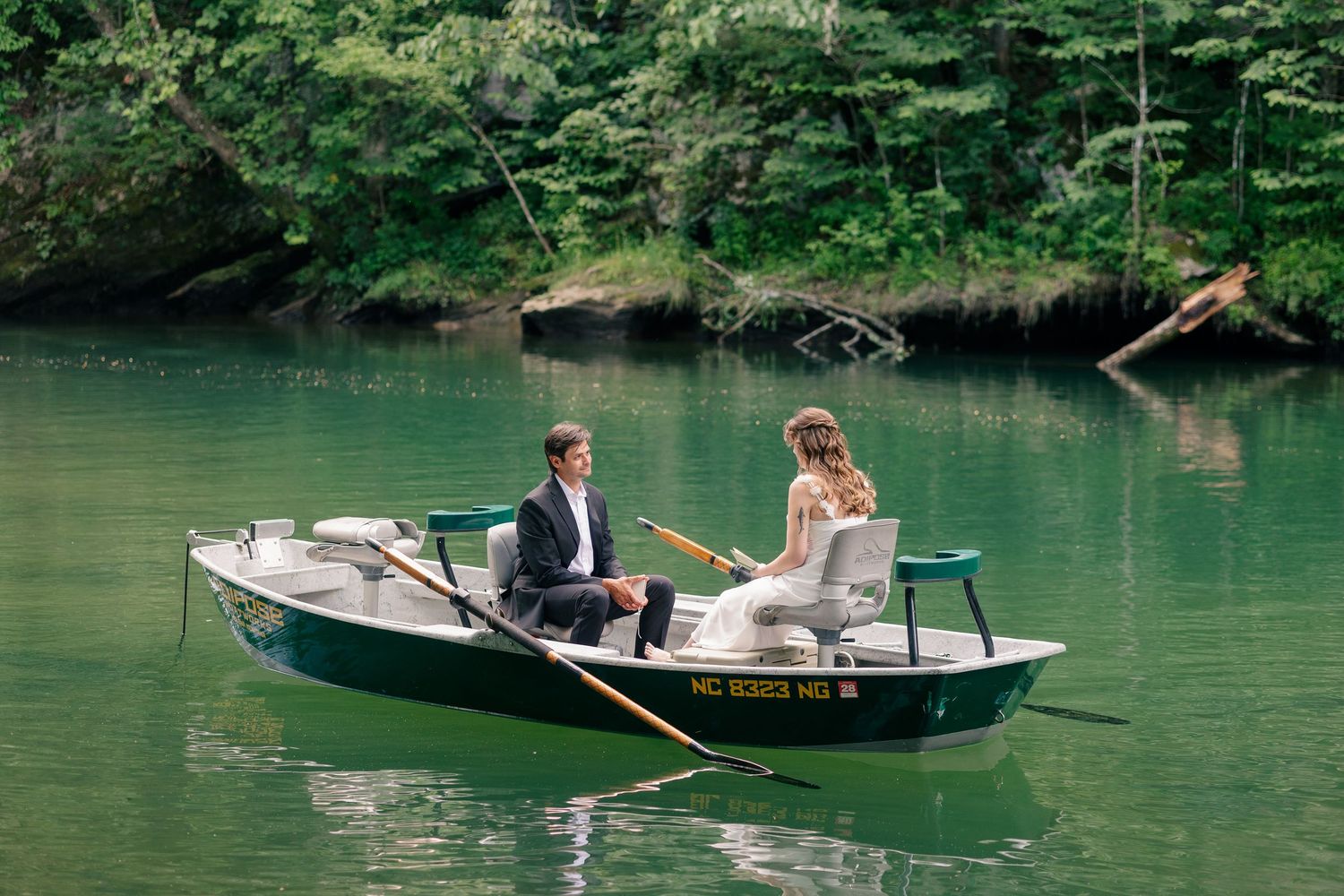 A couple in wedding attire rowing a green boat on a calm lake surrounded by lush green trees.