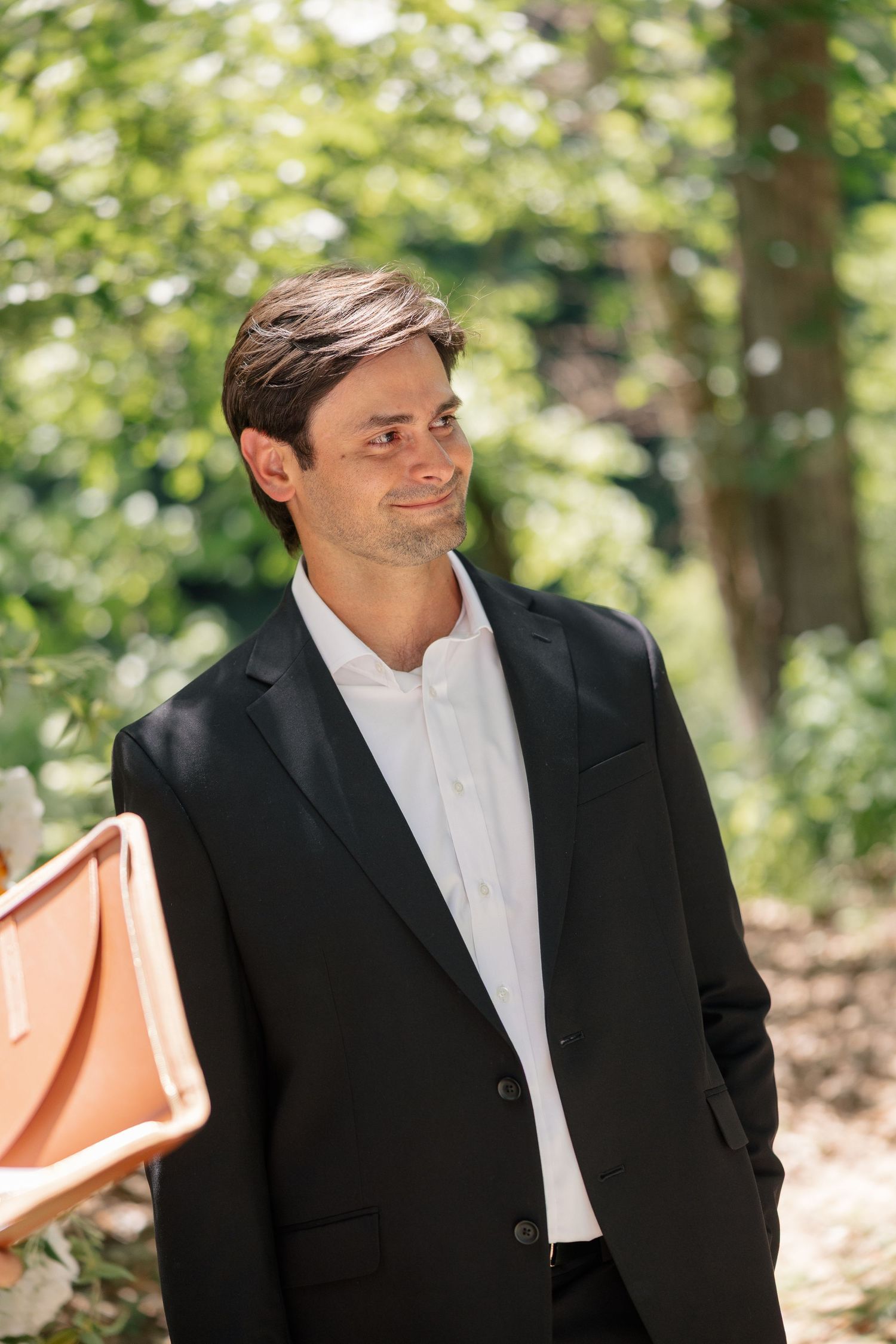 Handsome groom in black blazer smiles softly while standing outdoors in a sunlit green garden setting.