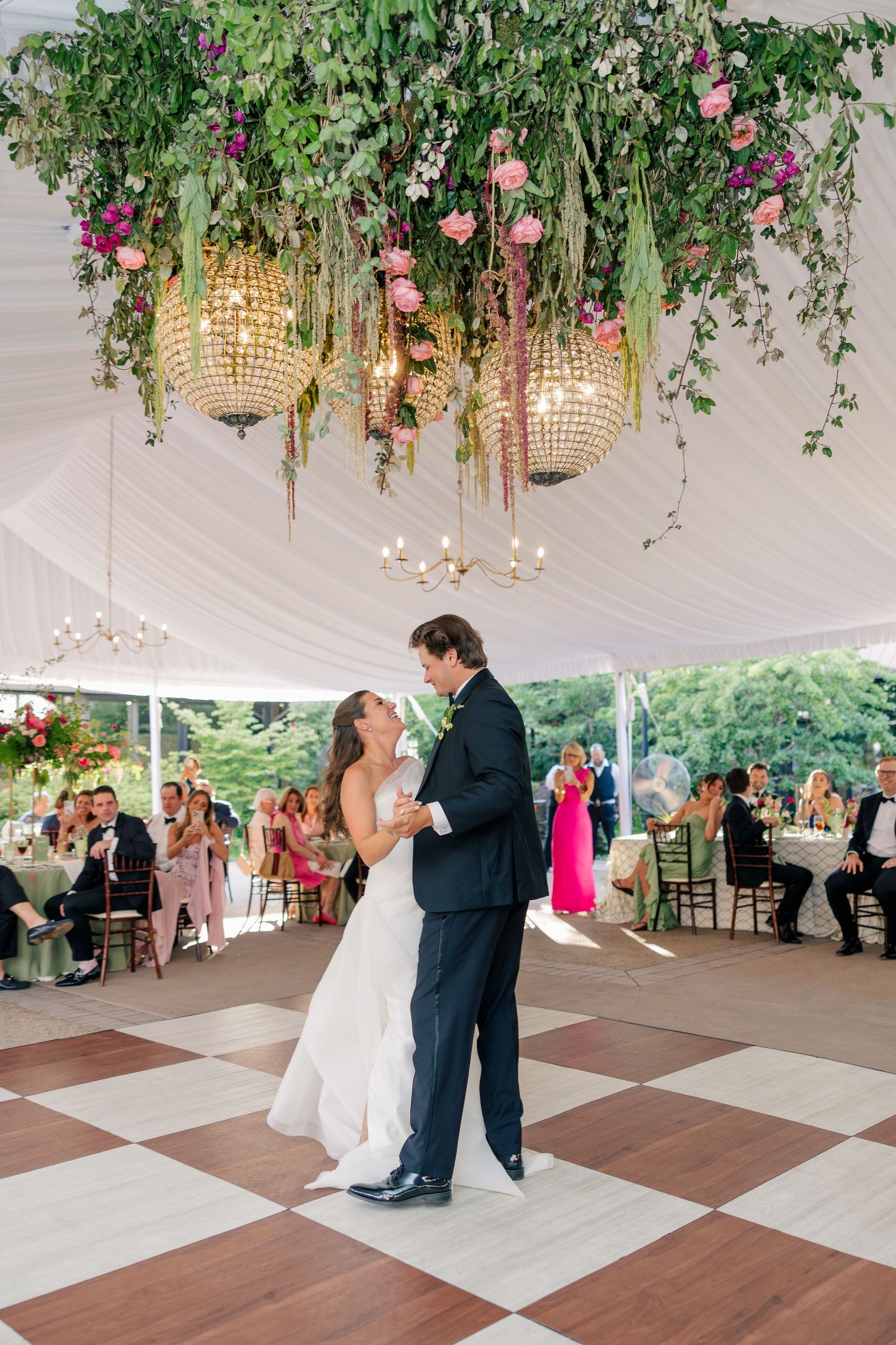 Bride and groom share first dance under stunning floral chandelier with pink roses in wedding tent.