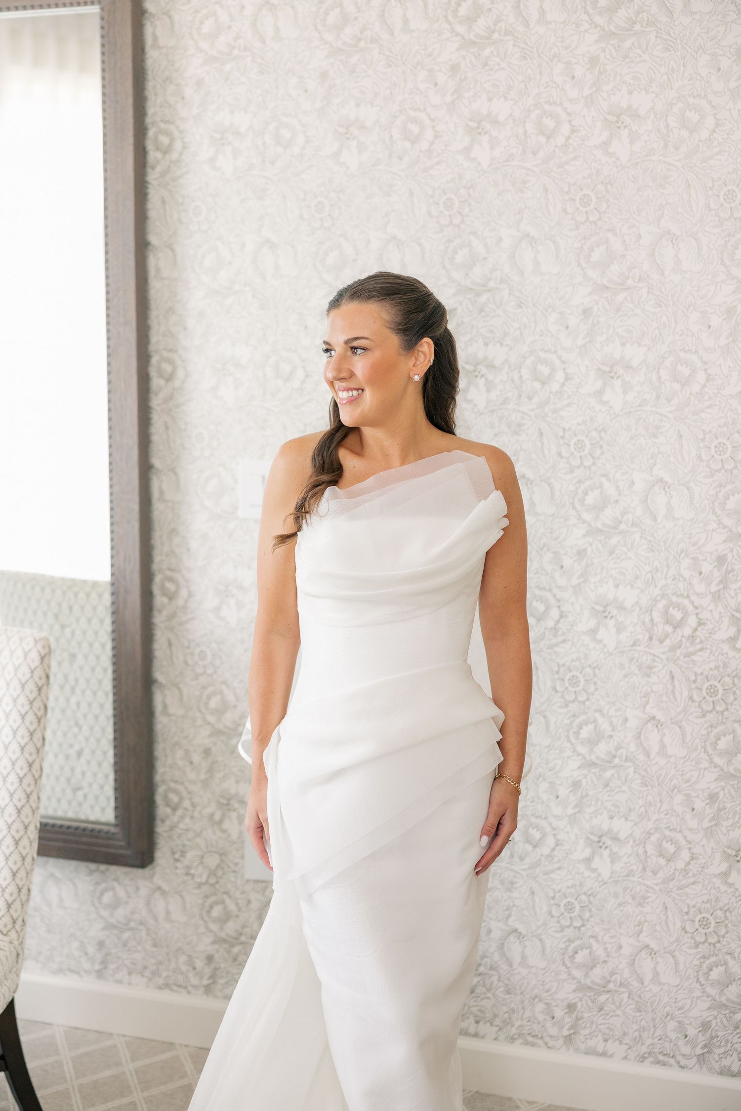 Smiling bride in a sleek white mermaid wedding gown posing in front of elegant grey patterned wallpaper.