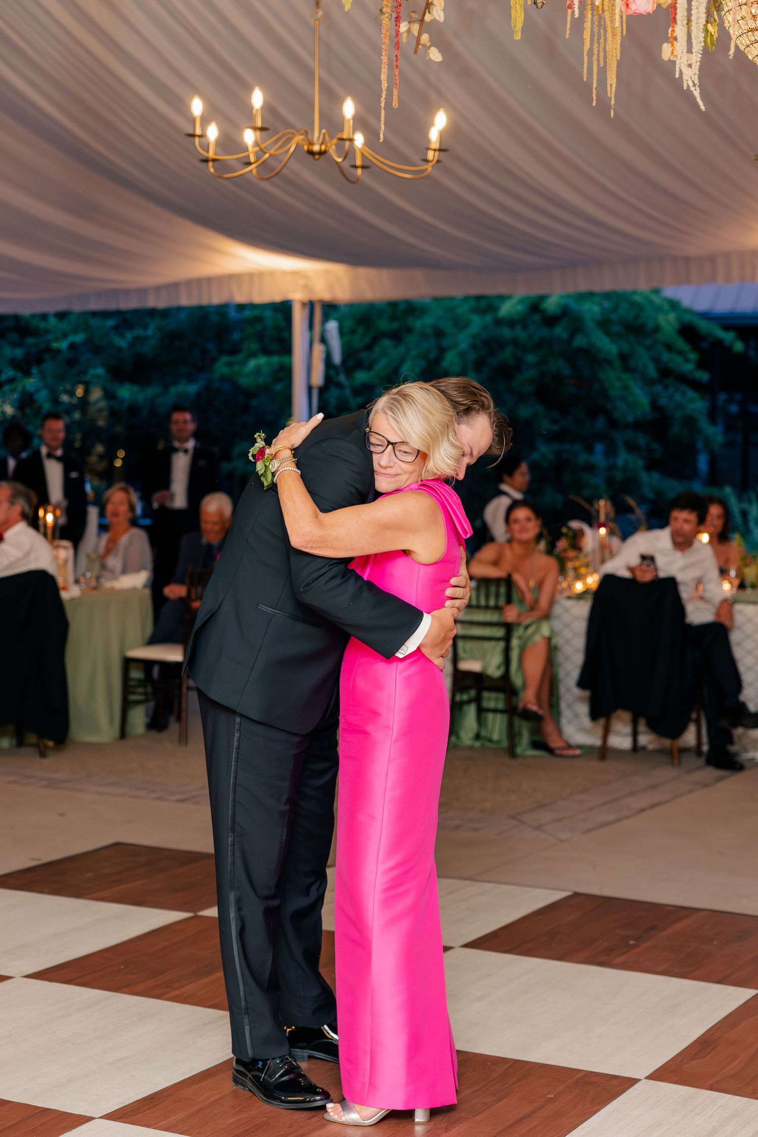 Groom shares an emotional dance with his mother in a pink gown under a chandelier at wedding reception.