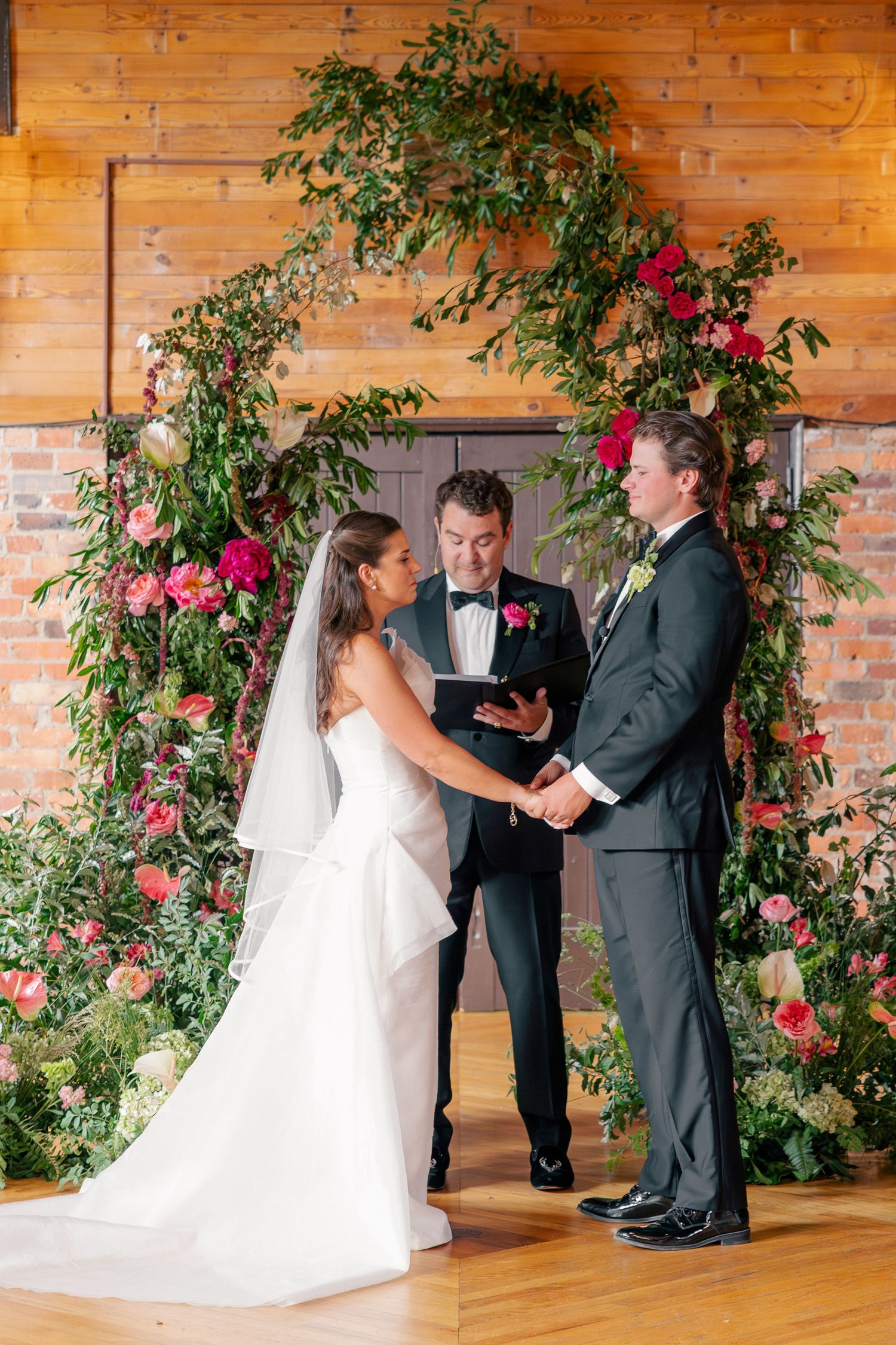 Bride and groom exchange vows beneath lush floral arch of pink roses and greenery at elegant wedding ceremony.