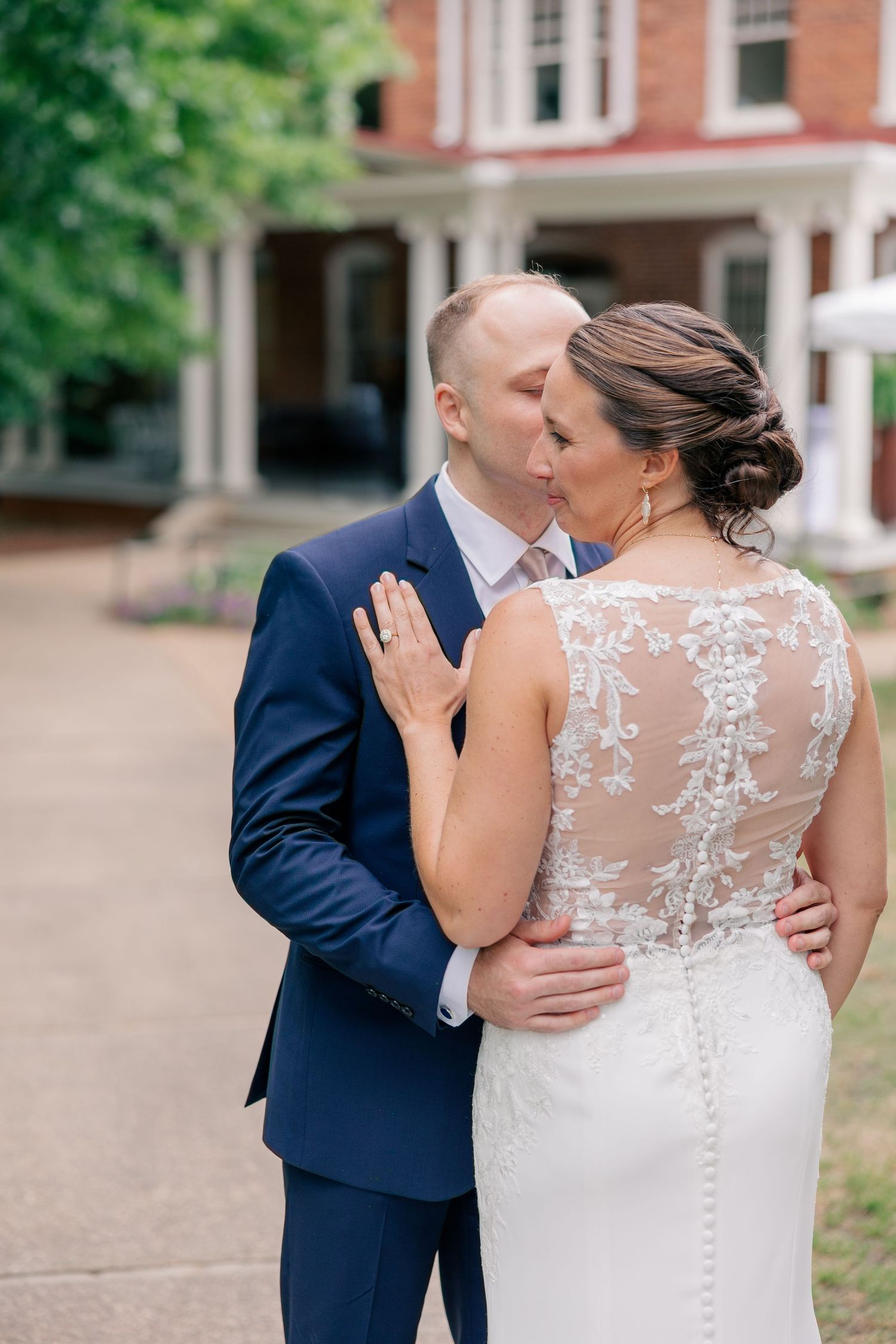 Groom in navy suit kisses bride in lace back wedding gown outdoors near brick manor house.