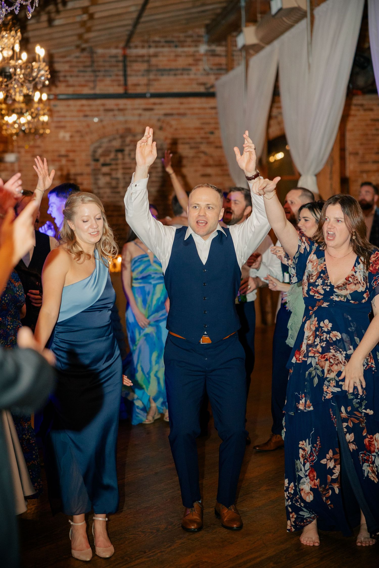 Energetic wedding guest raises hands on dance floor surrounded by celebrating guests in brick venue.