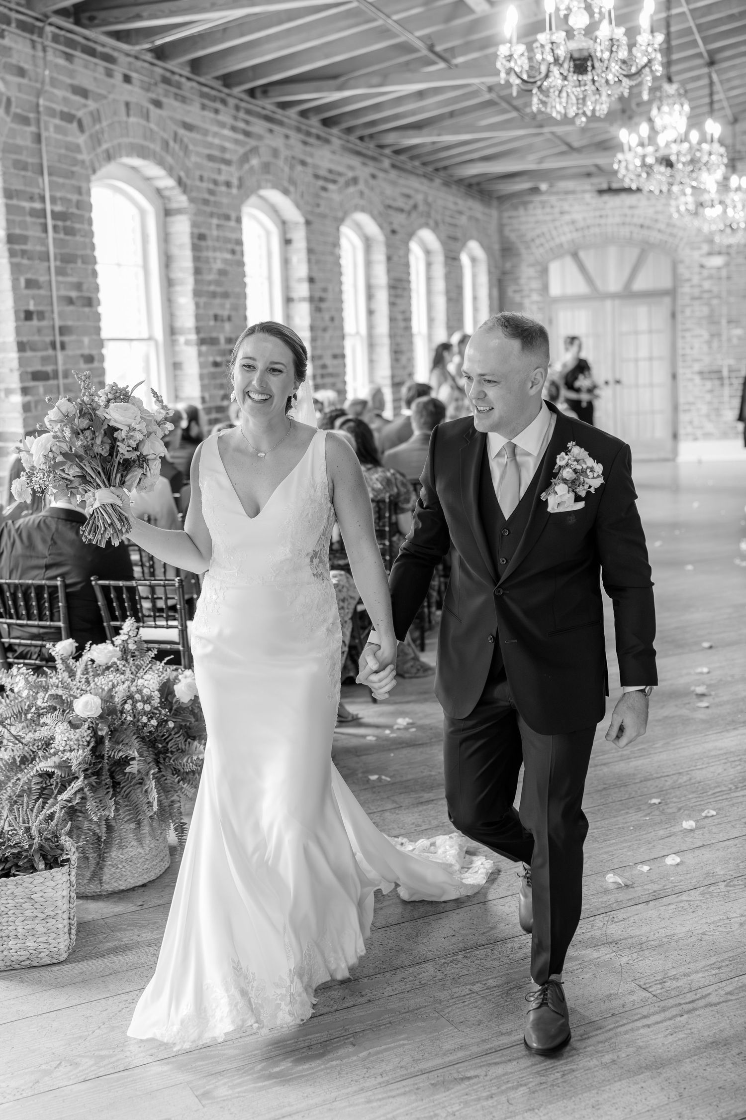 Bride and groom walk joyfully down the aisle in a rustic brick wedding venue with chandeliers.
