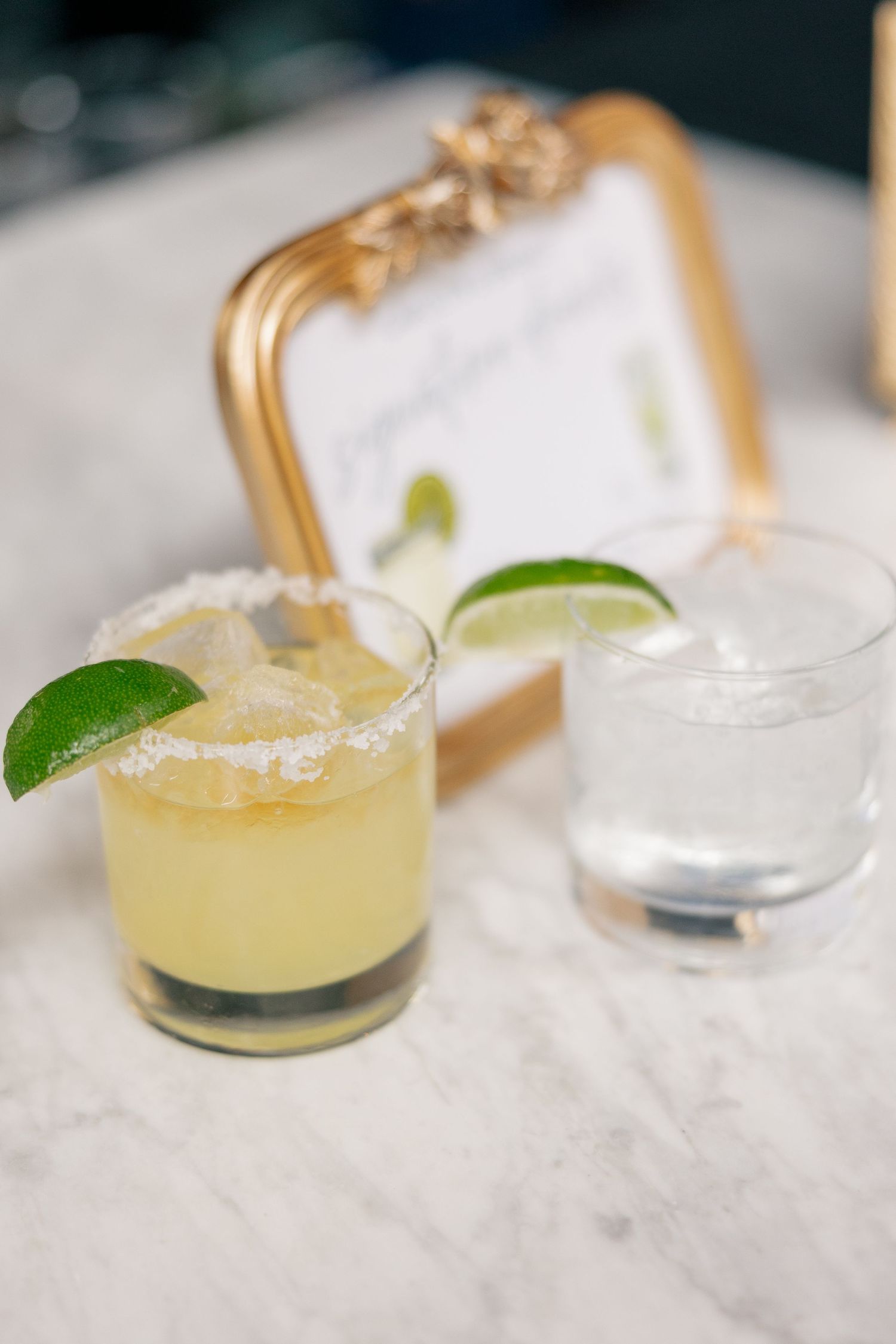 Two signature wedding cocktails with lime garnish and salted rim displayed on marble surface with gold frame.