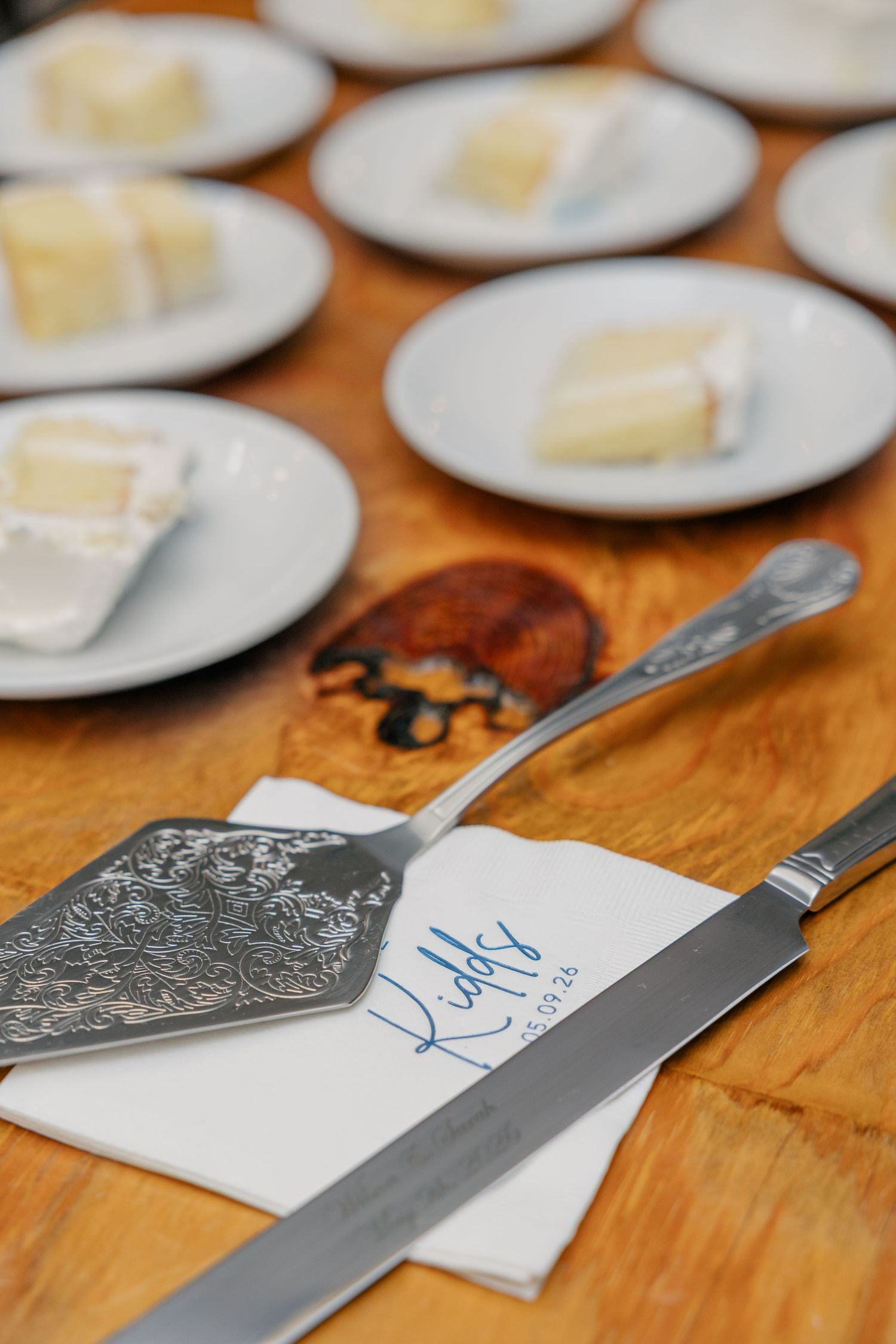 Elegant wedding cake server set rests on personalized napkin beside plated white cake slices on wood table.