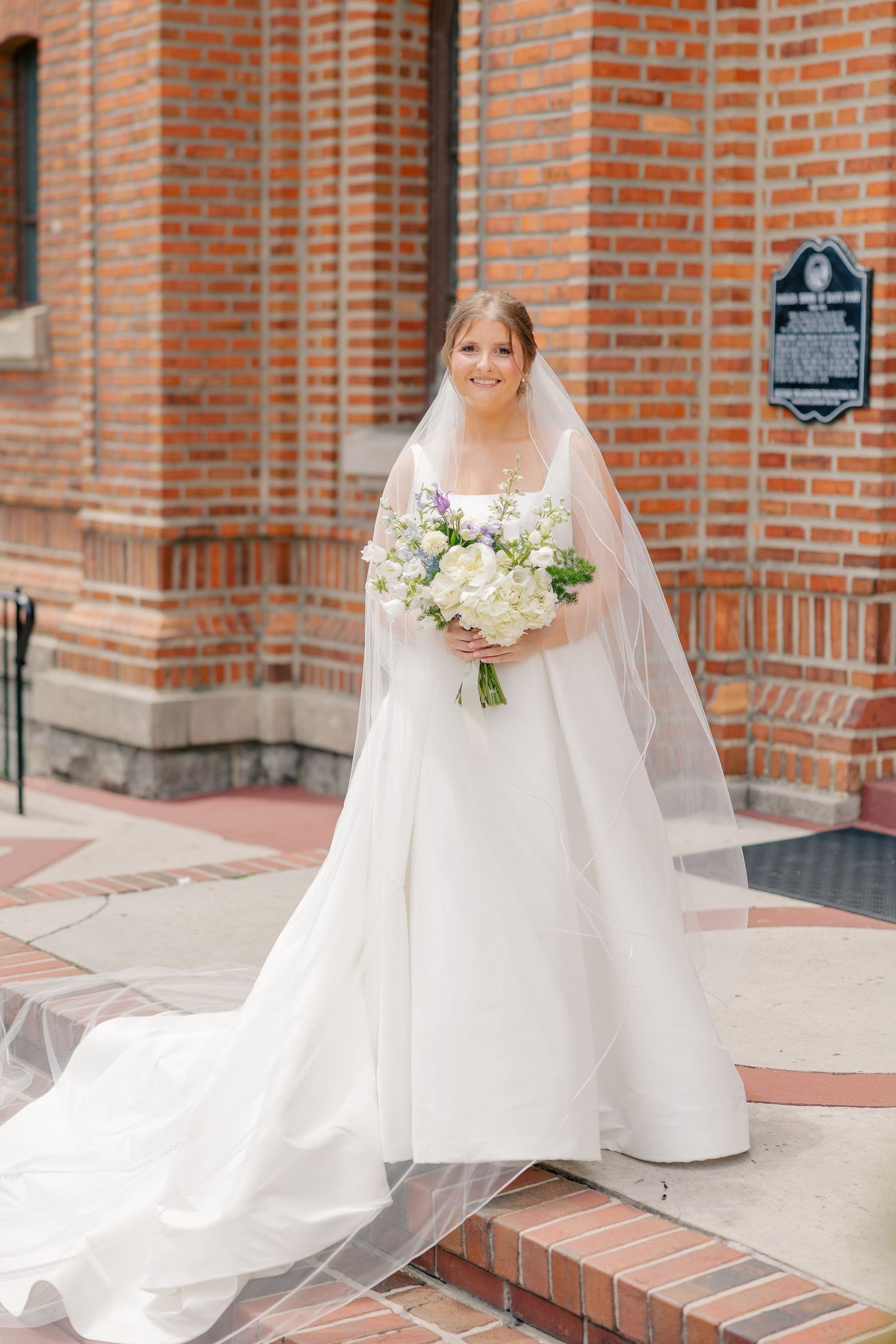 Bride in elegant white ball gown with cathedral veil holds white floral bouquet while posing outside brick church.