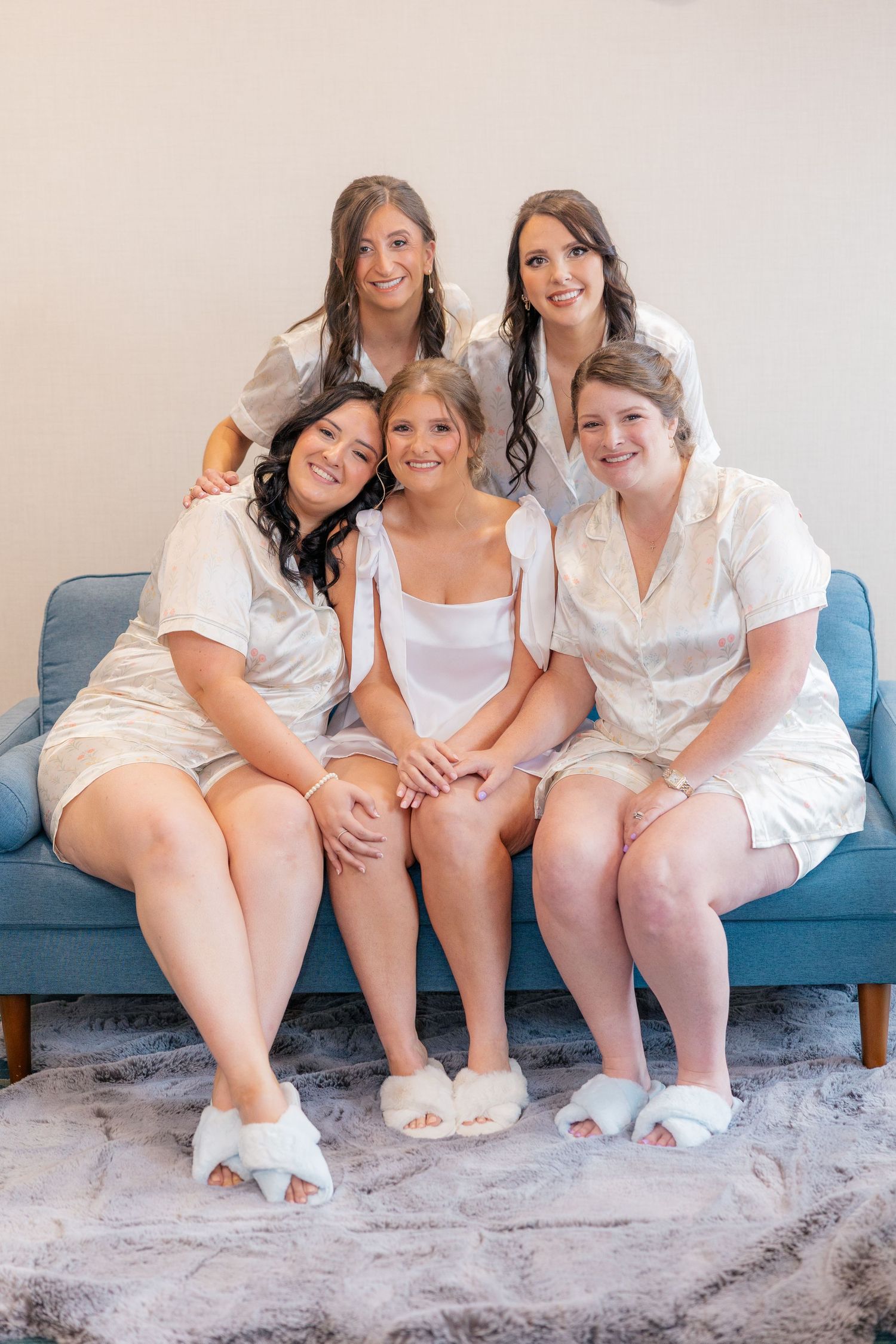 Bride and four bridesmaids pose together in white satin robes and fluffy slippers on a blue couch.