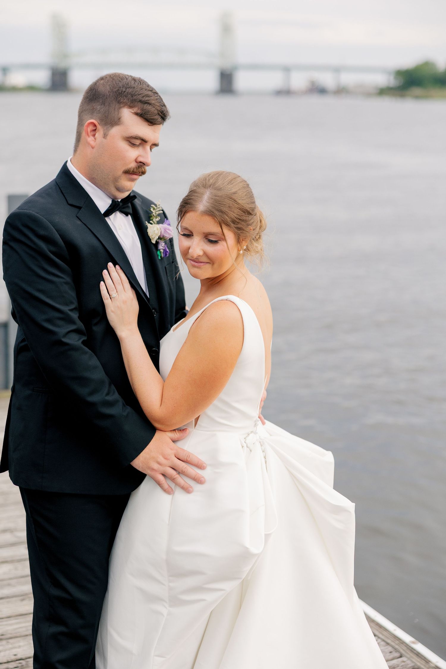 Bride and groom share an intimate embrace on a waterfront dock with a bridge in the background.