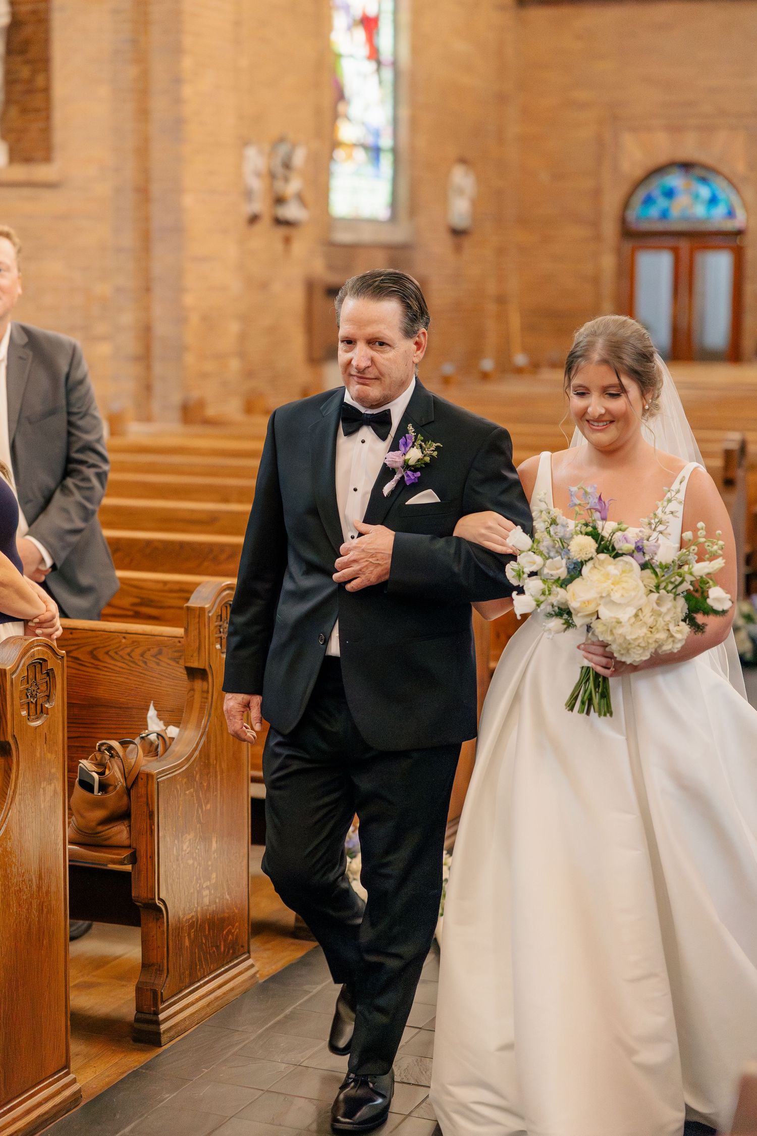 Bride walks down church aisle with escort, holding white and blue floral bouquet, smiling joyfully.