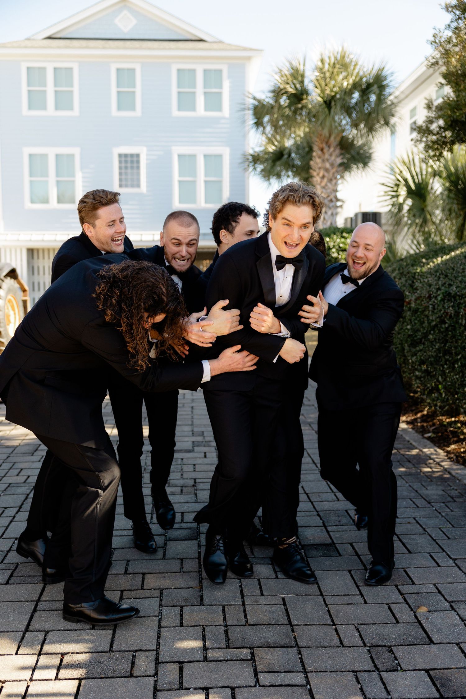 A group in formal attire playfully restrains a groomsman in a tuxedo on a brick patio outside a blue house.